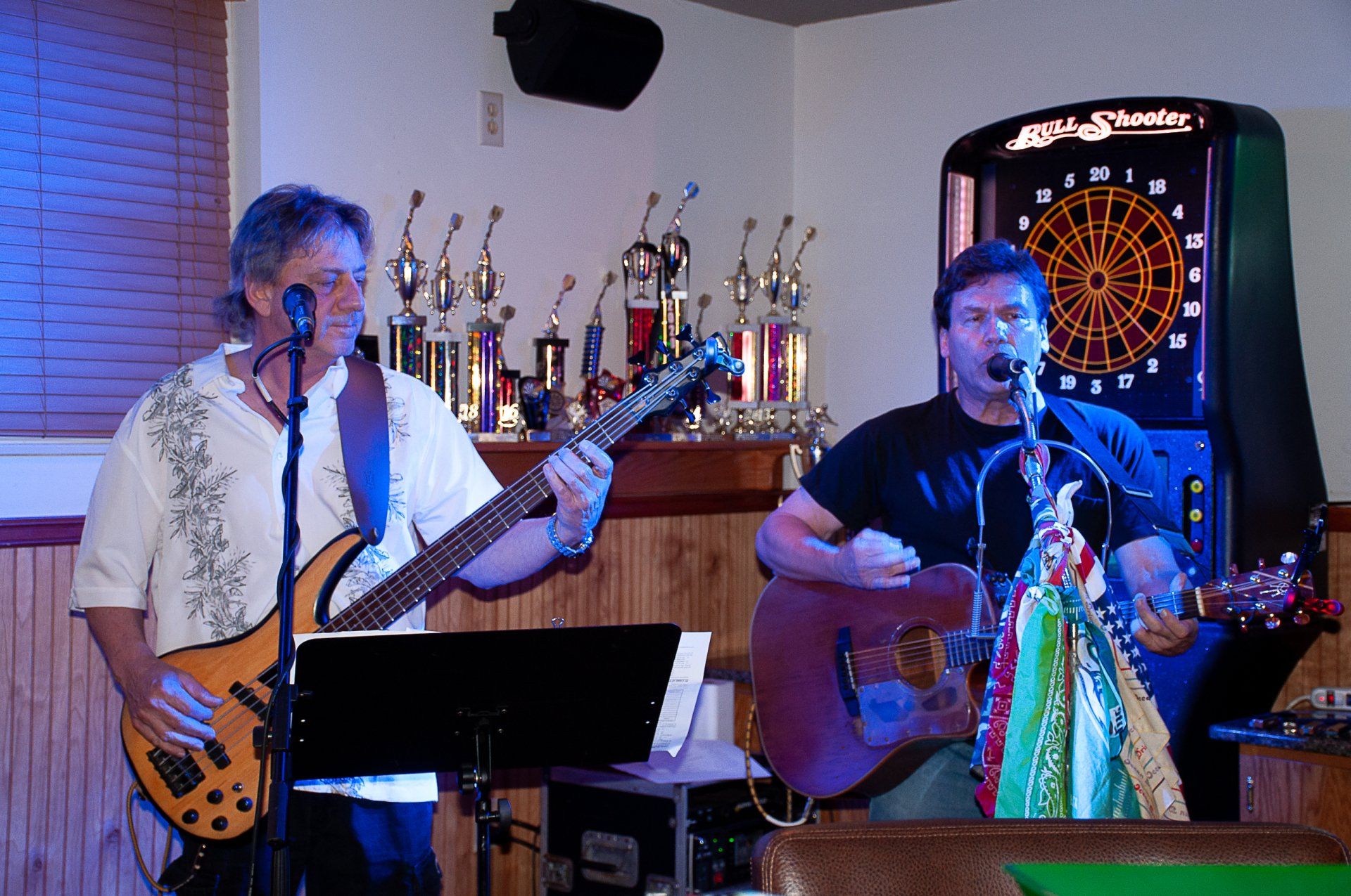 Two musicians performing live on stage, one with bass guitar and one with acoustic guitar, near a dartboard.