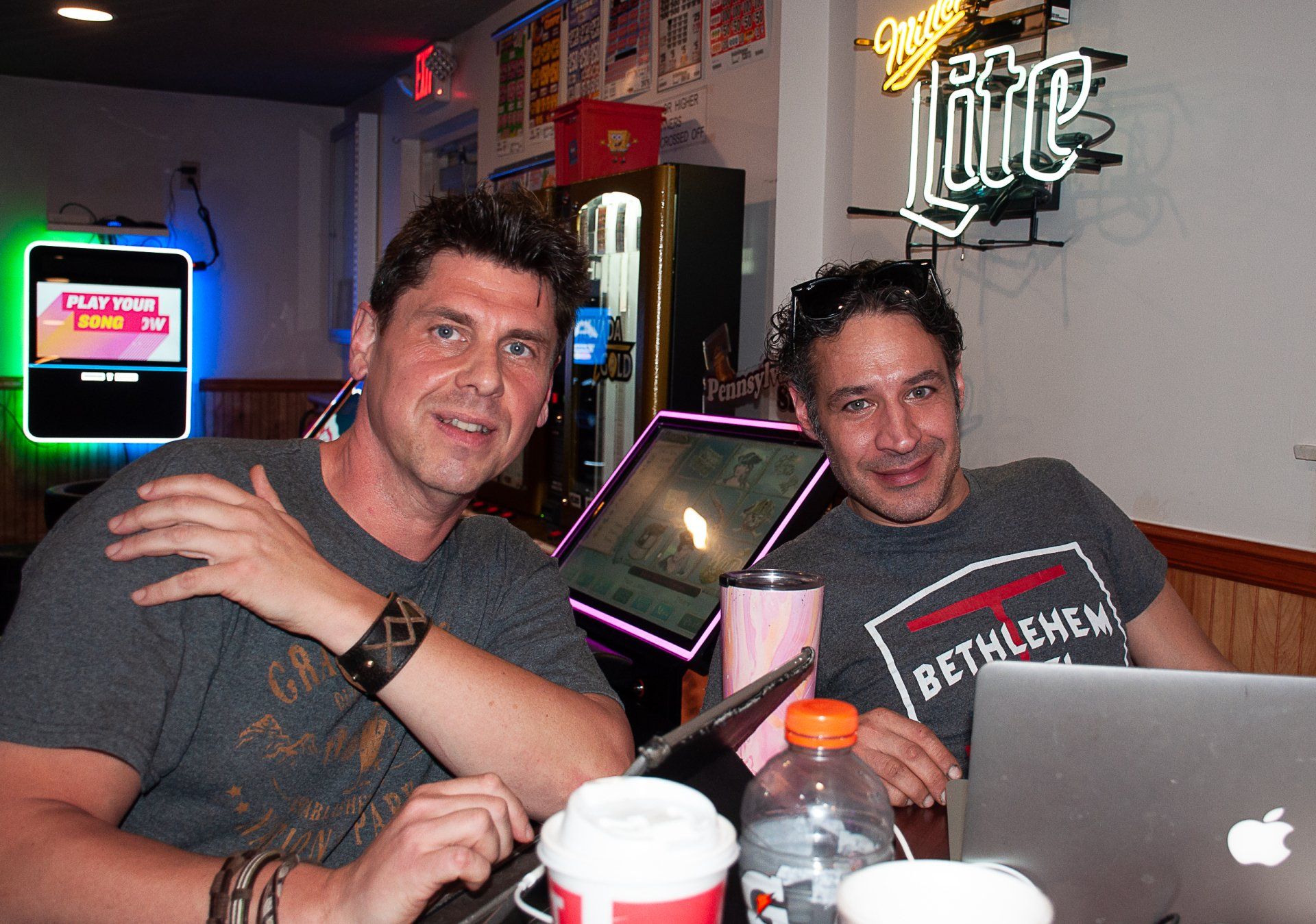 Two men seated at a table in a neon-lit bar, smiling beside drinks and a laptop.
