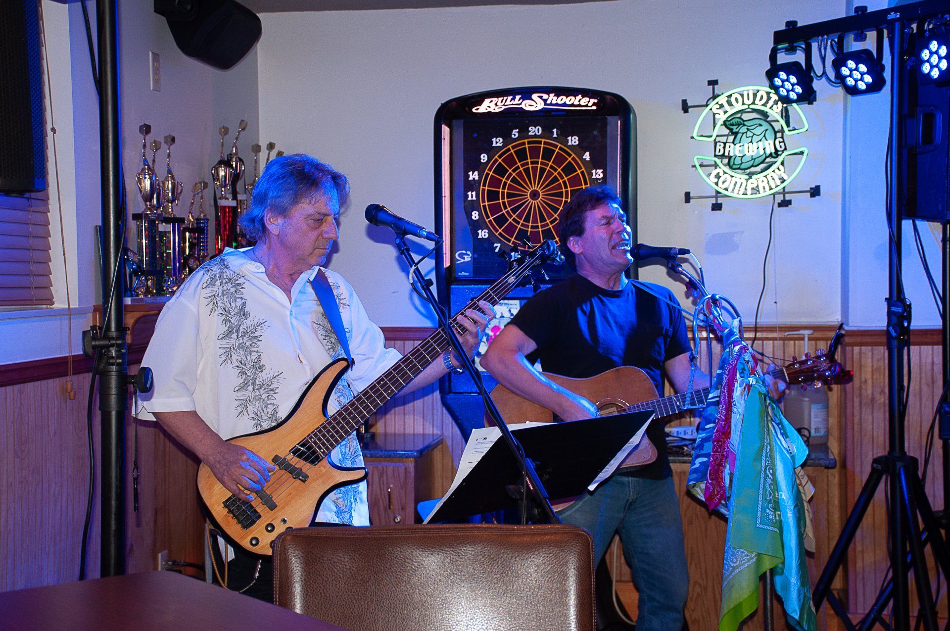Two musicians perform on stage with guitars under blue and purple lights, with dartboards on the wall.