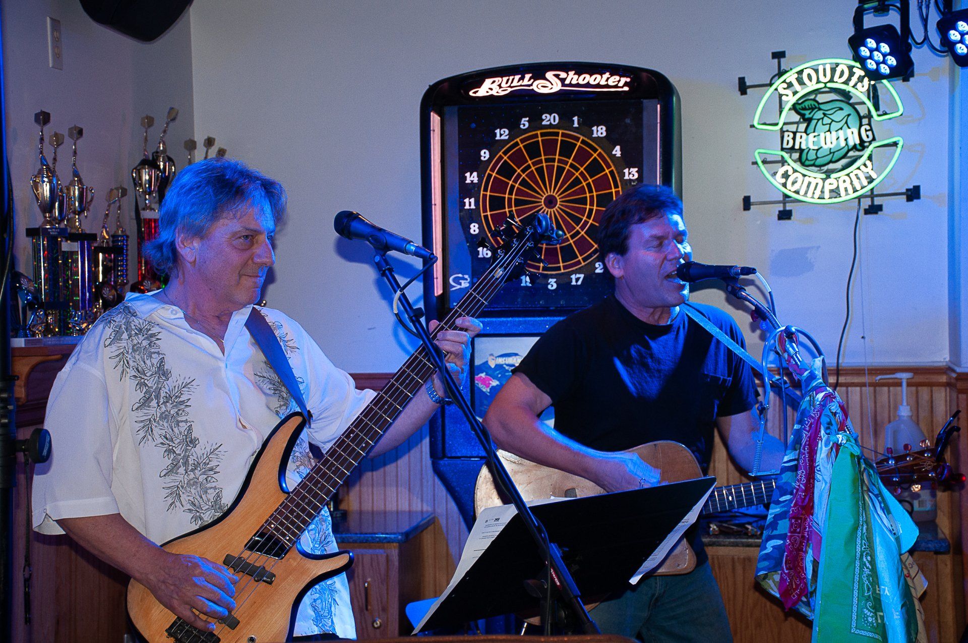 Two musicians performing onstage, one playing guitar and one singing into a microphone, with a dartboard behind them.