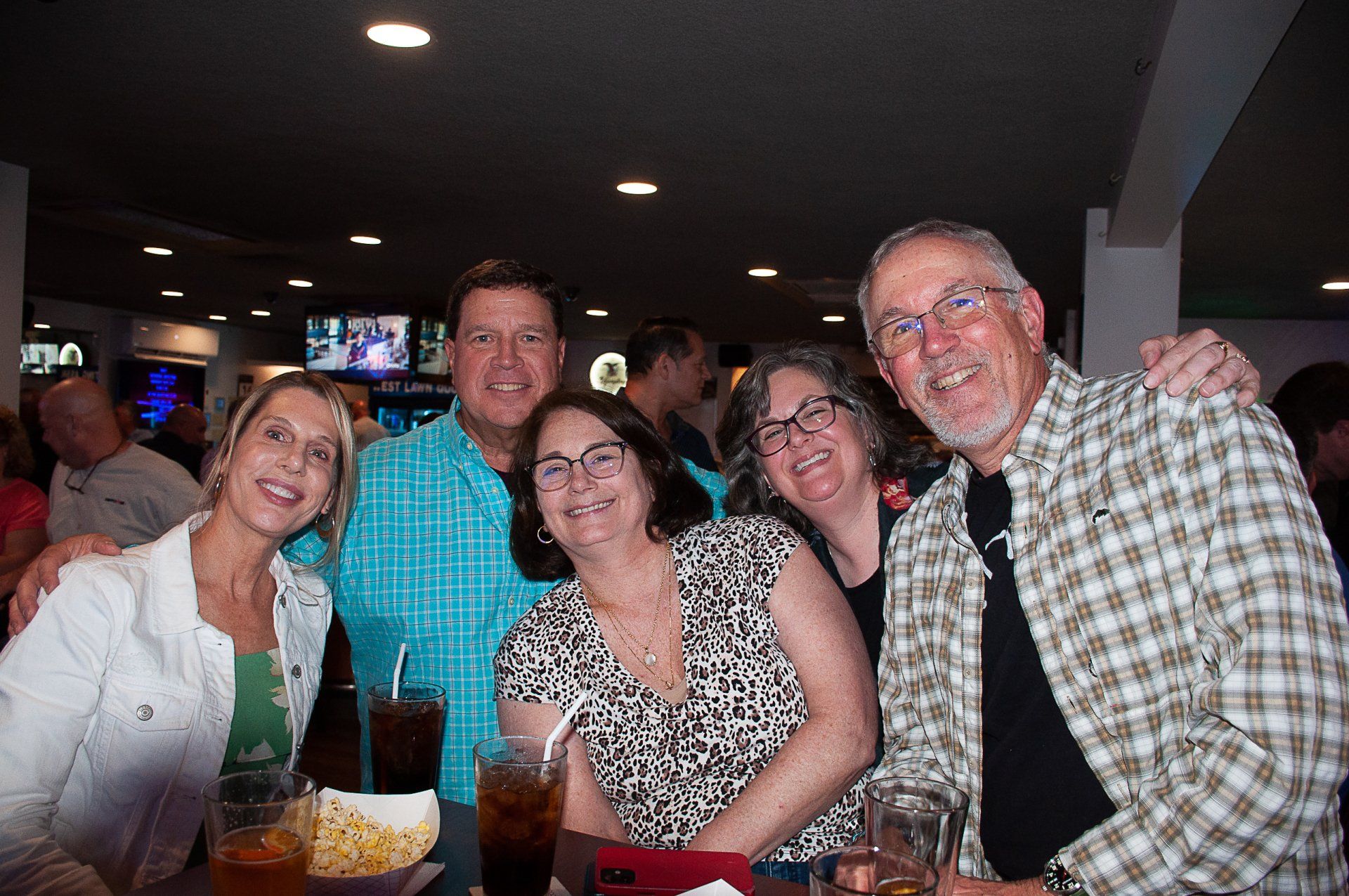 Five smiling people posing at a dimly lit restaurant table with drinks and desserts