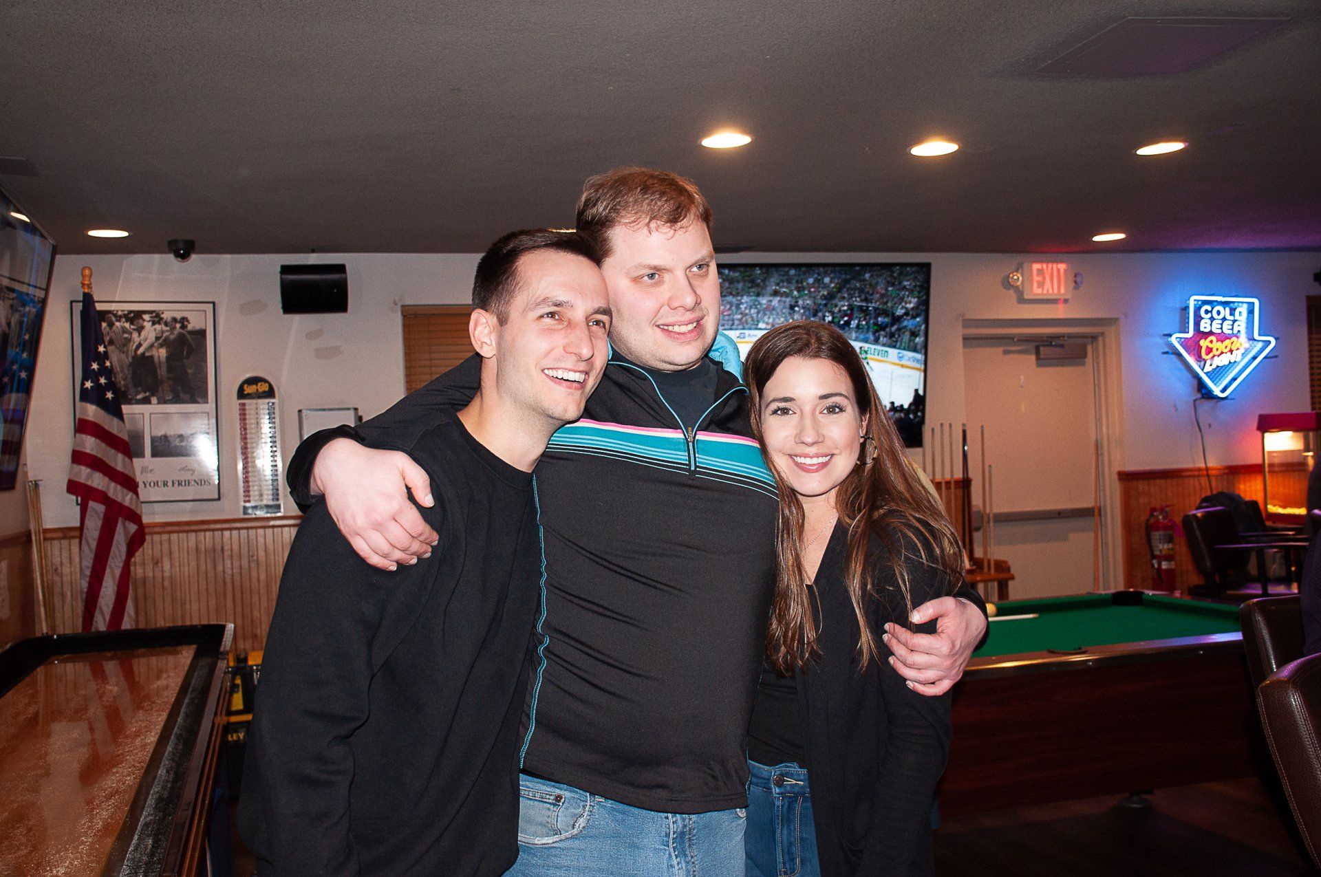 Three people smiling and hugging in a pool hall with an American flag and neon signs.