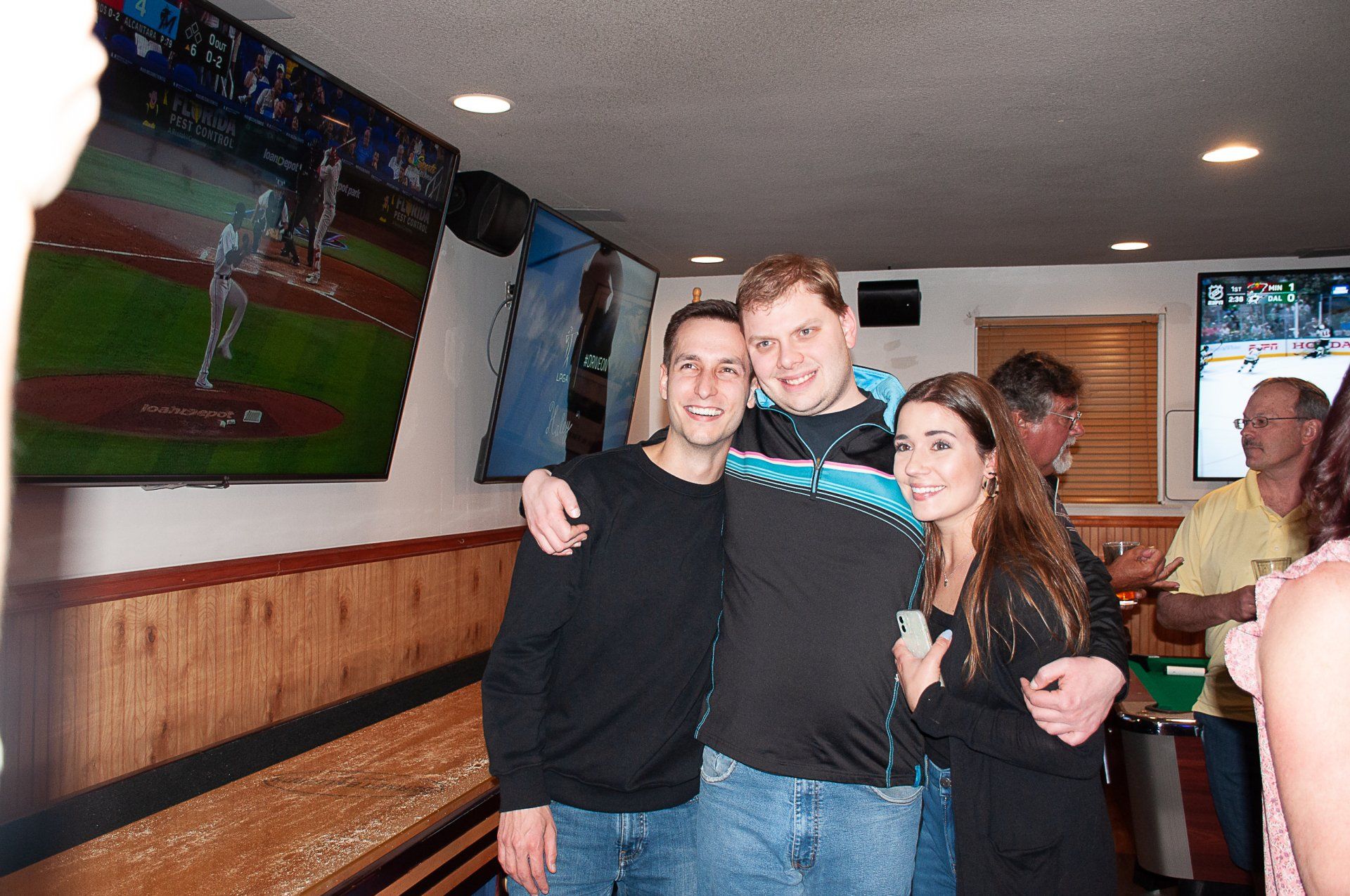 Group posing together in a sports bar, smiling with arms around each other, TV screens in the background