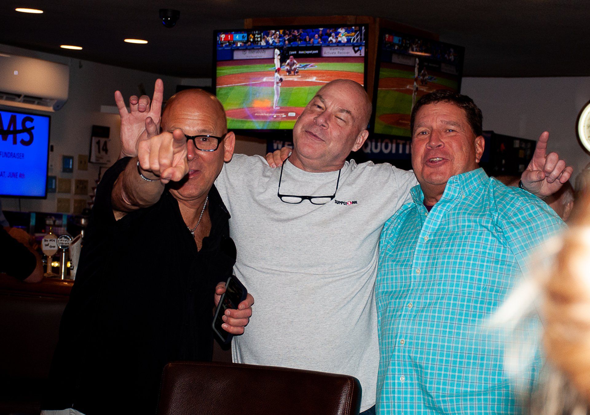 Three men smiling and posing in a dimly lit bar, with a TV showing a sports game in the background