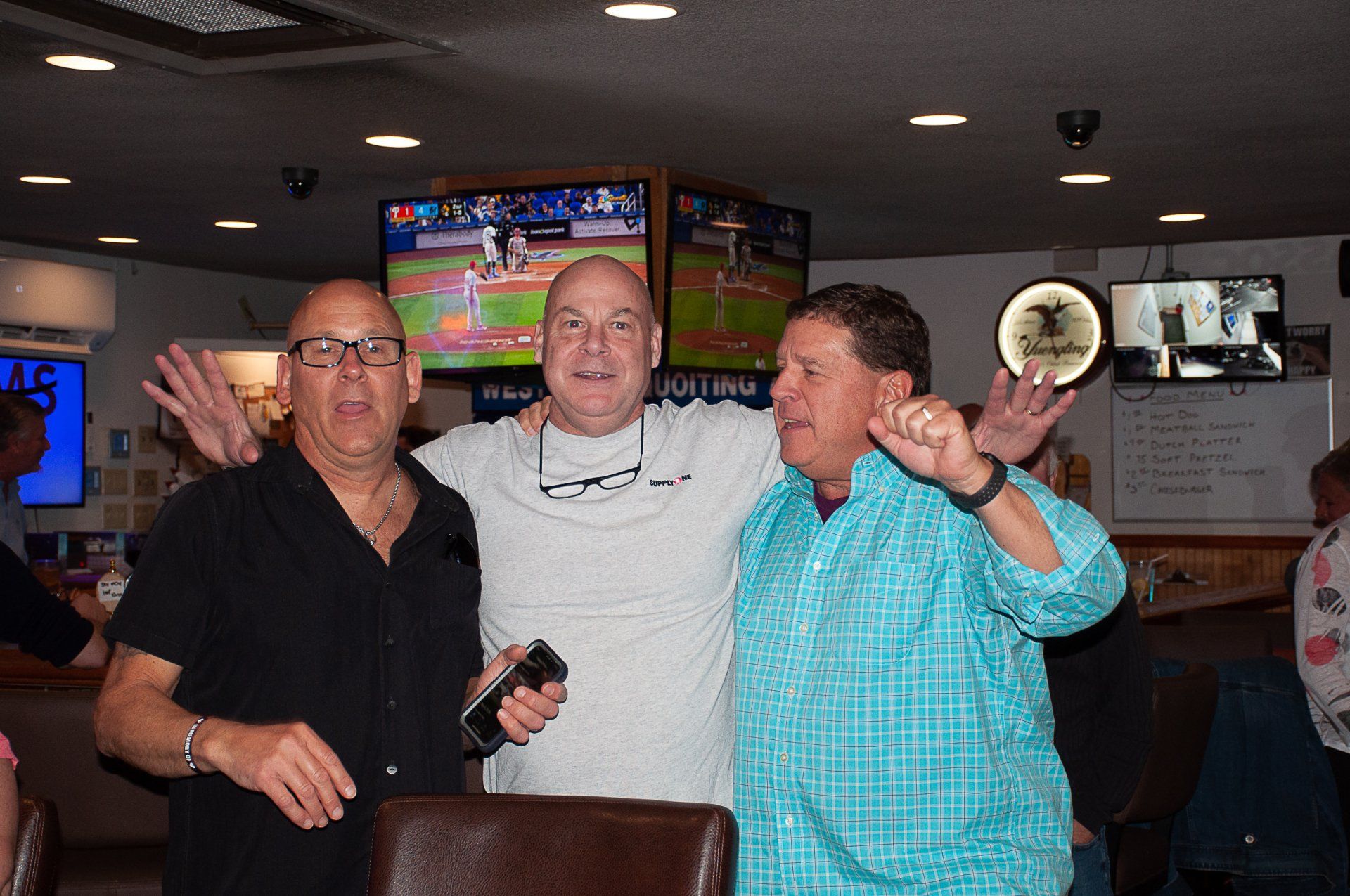 Three people smiling and posing in a bar, with TVs and drinks in the background.