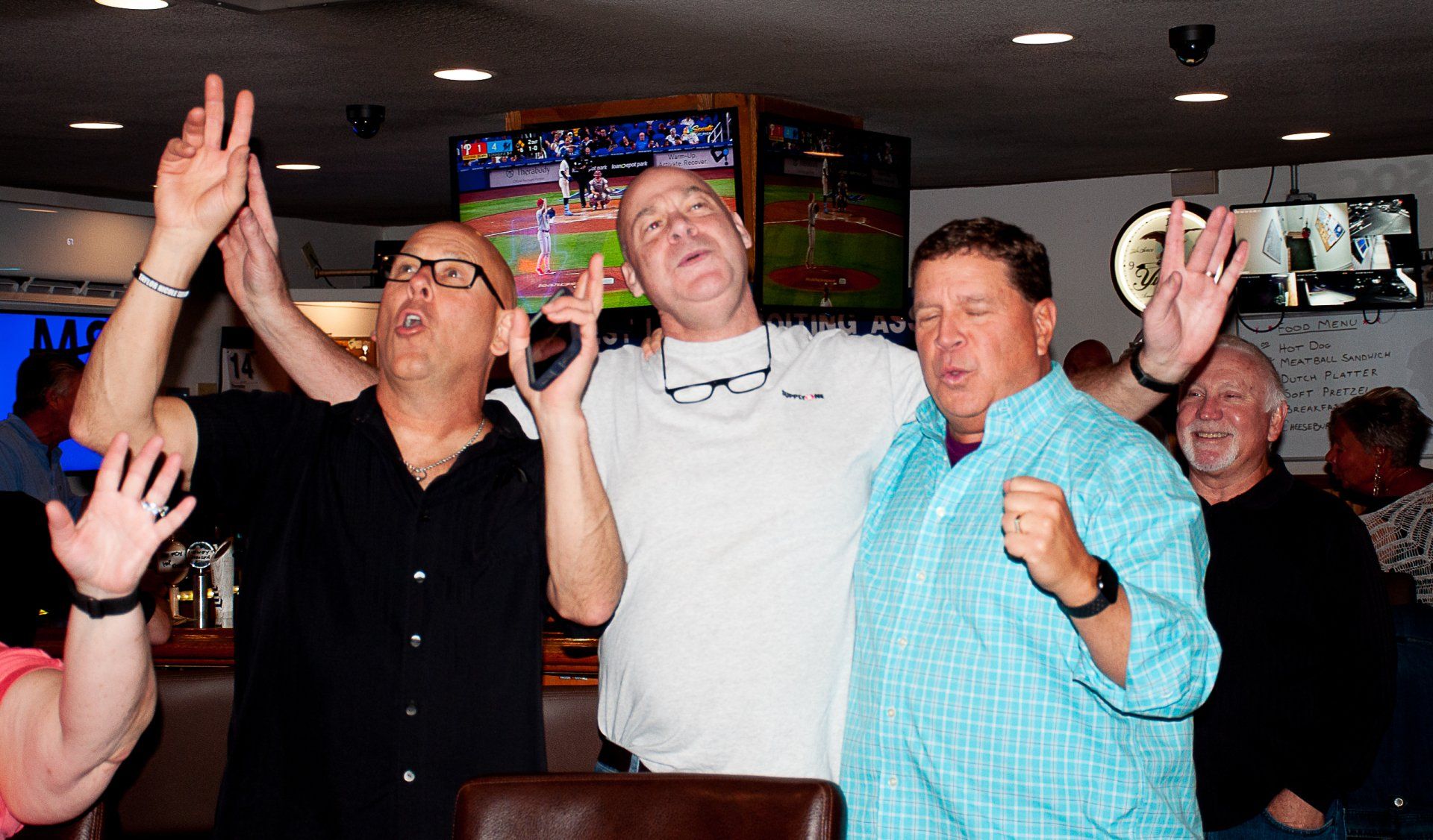 Three men cheering with raised arms in a sports bar, with a TV showing a game in the background