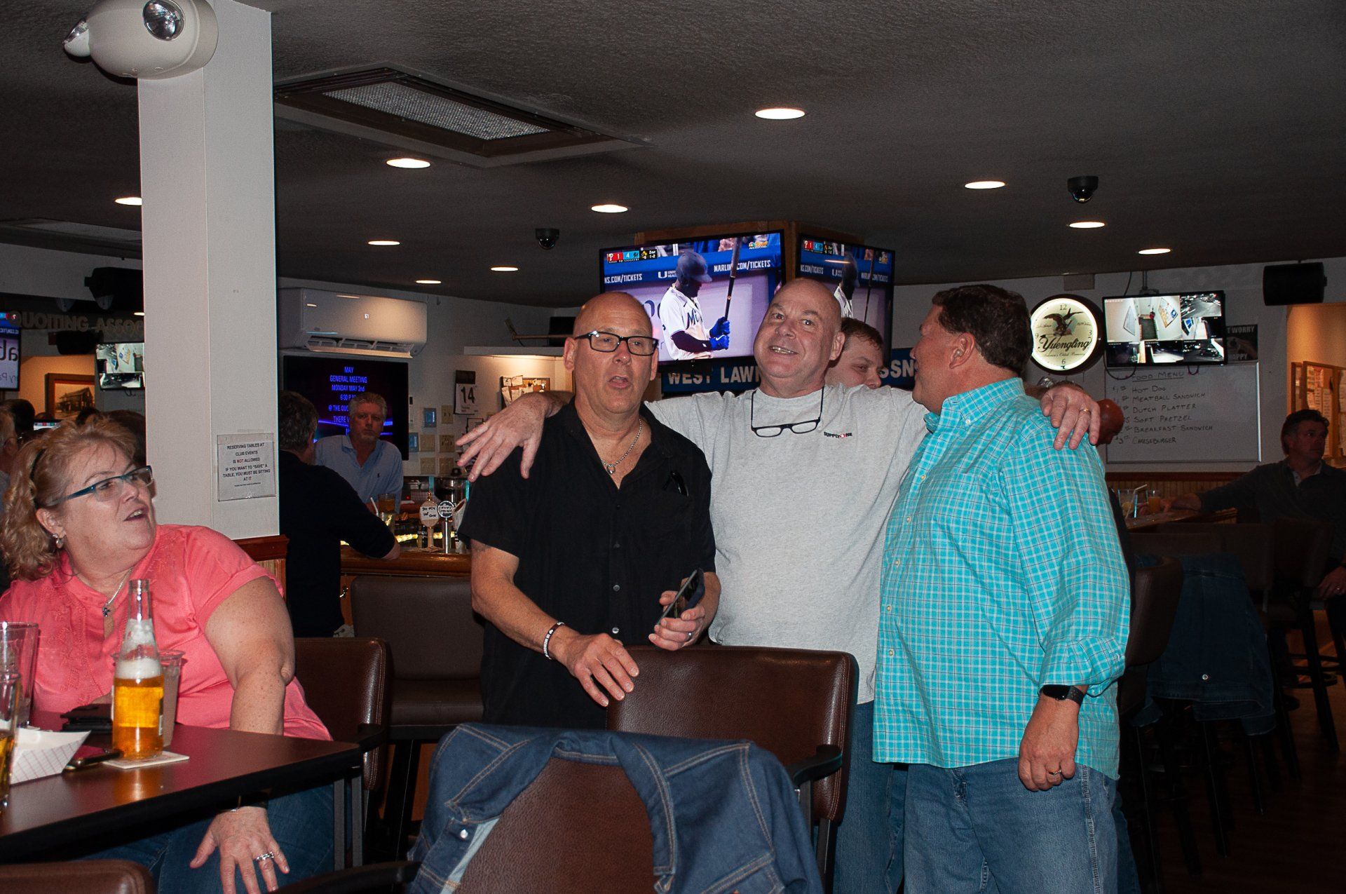 People posing together in a dimly lit bar, with TVs and other patrons in the background