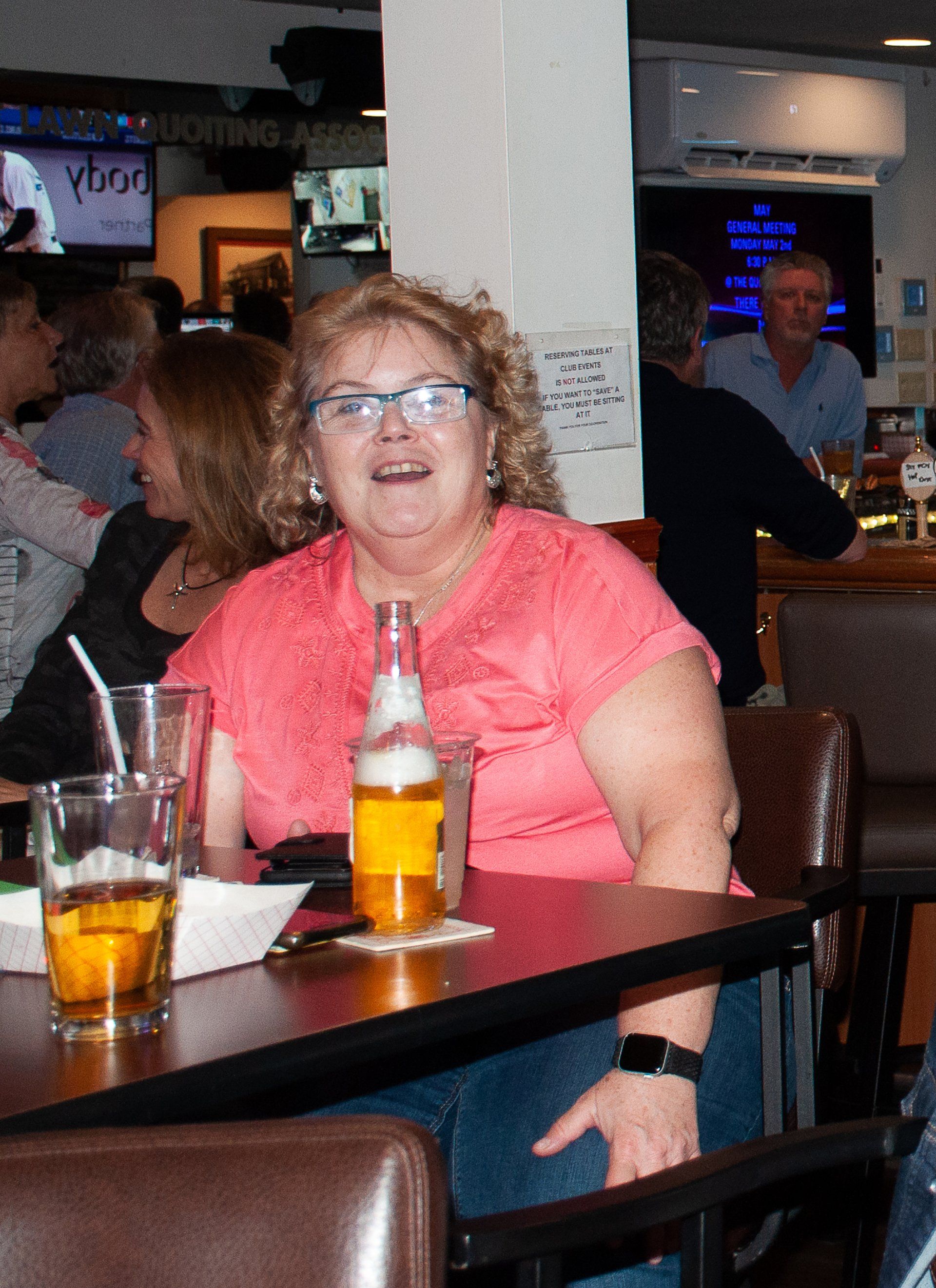 Woman in coral shirt sitting at a bar table with drinks, smiling in a dimly lit restaurant.