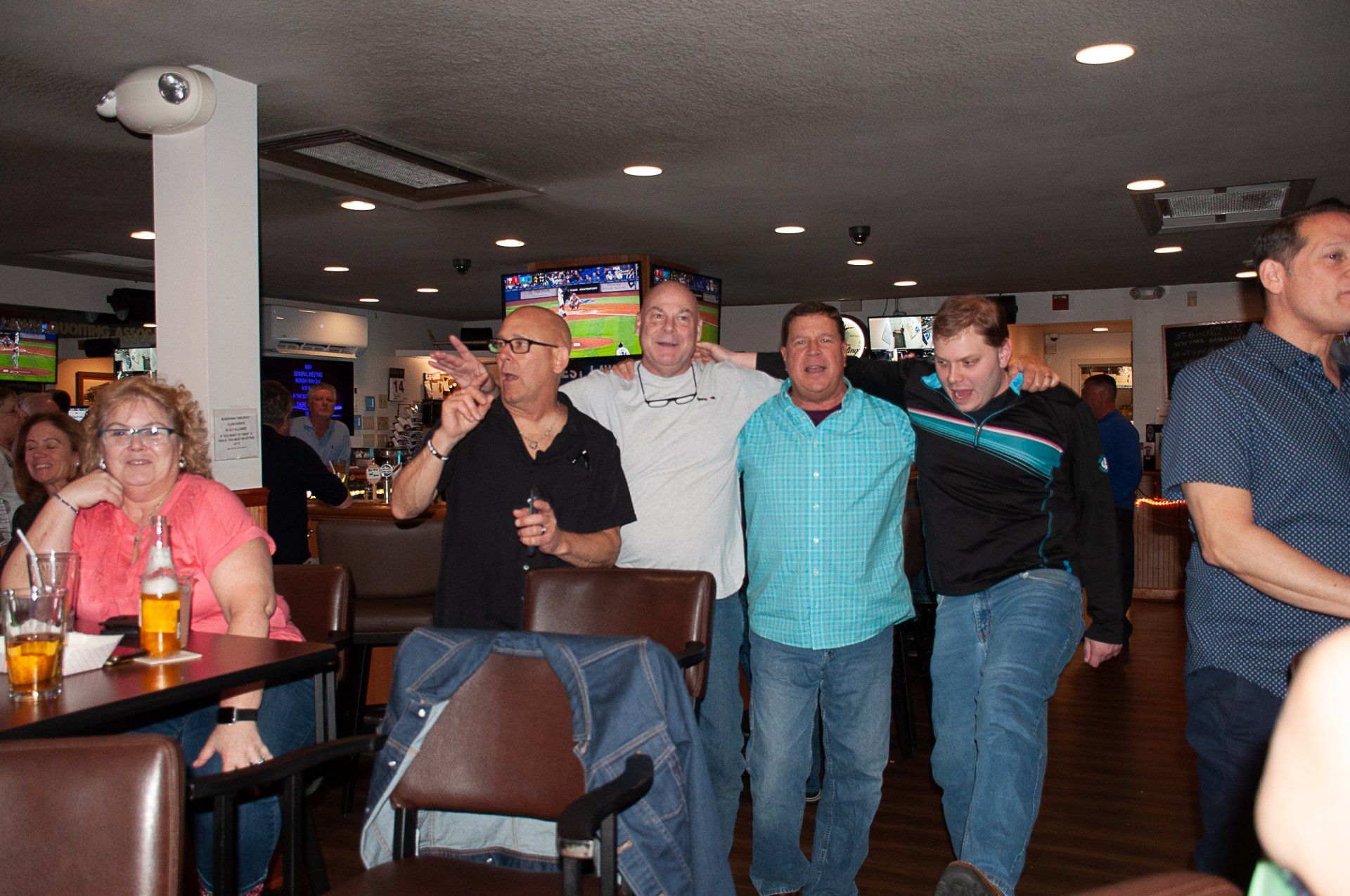 Group of people standing and sitting in a dimly lit restaurant or bar, watching something off-camera.