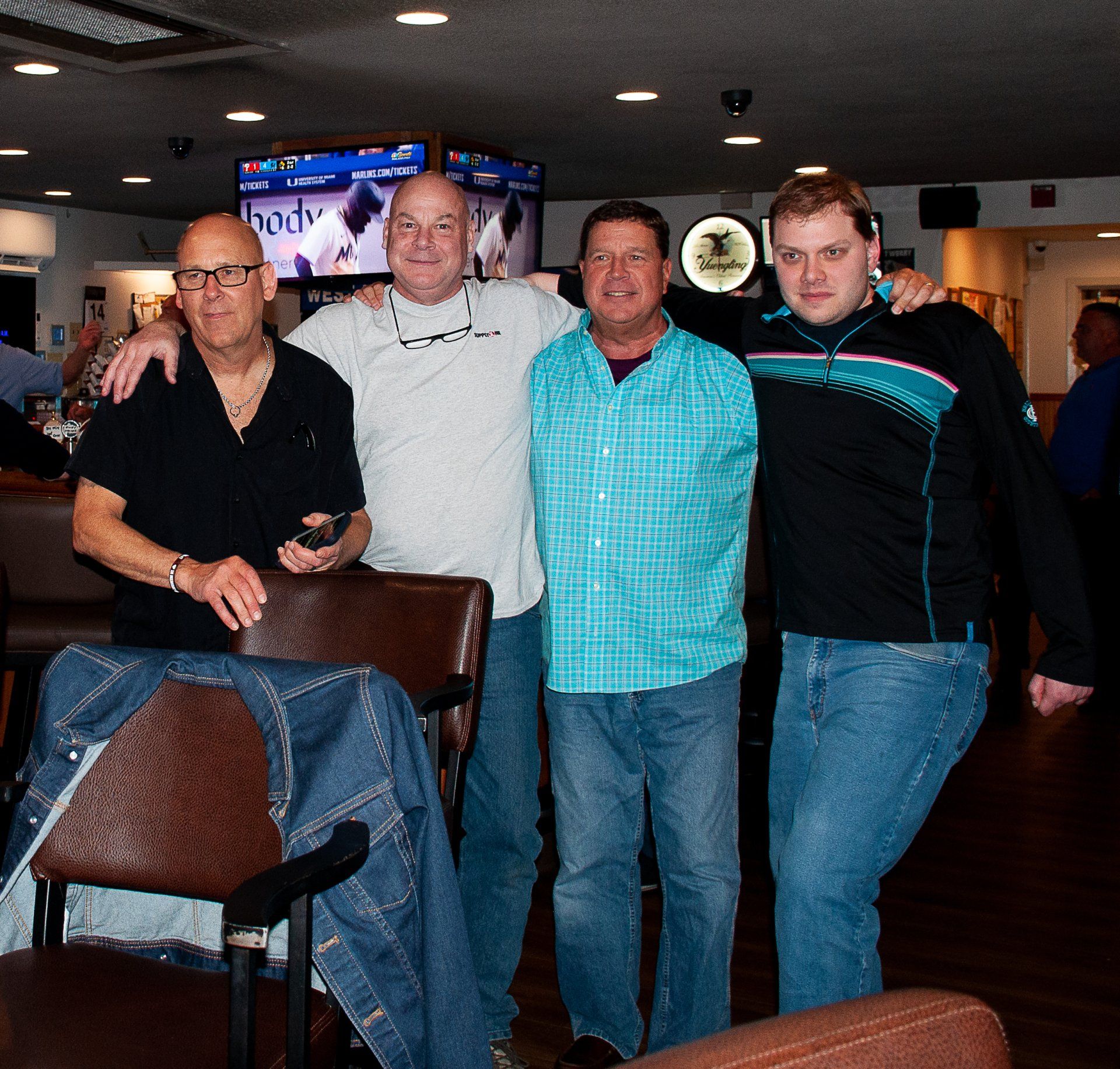 Four men posing together in a dimly lit bar, with one leaning on a chair in the foreground.