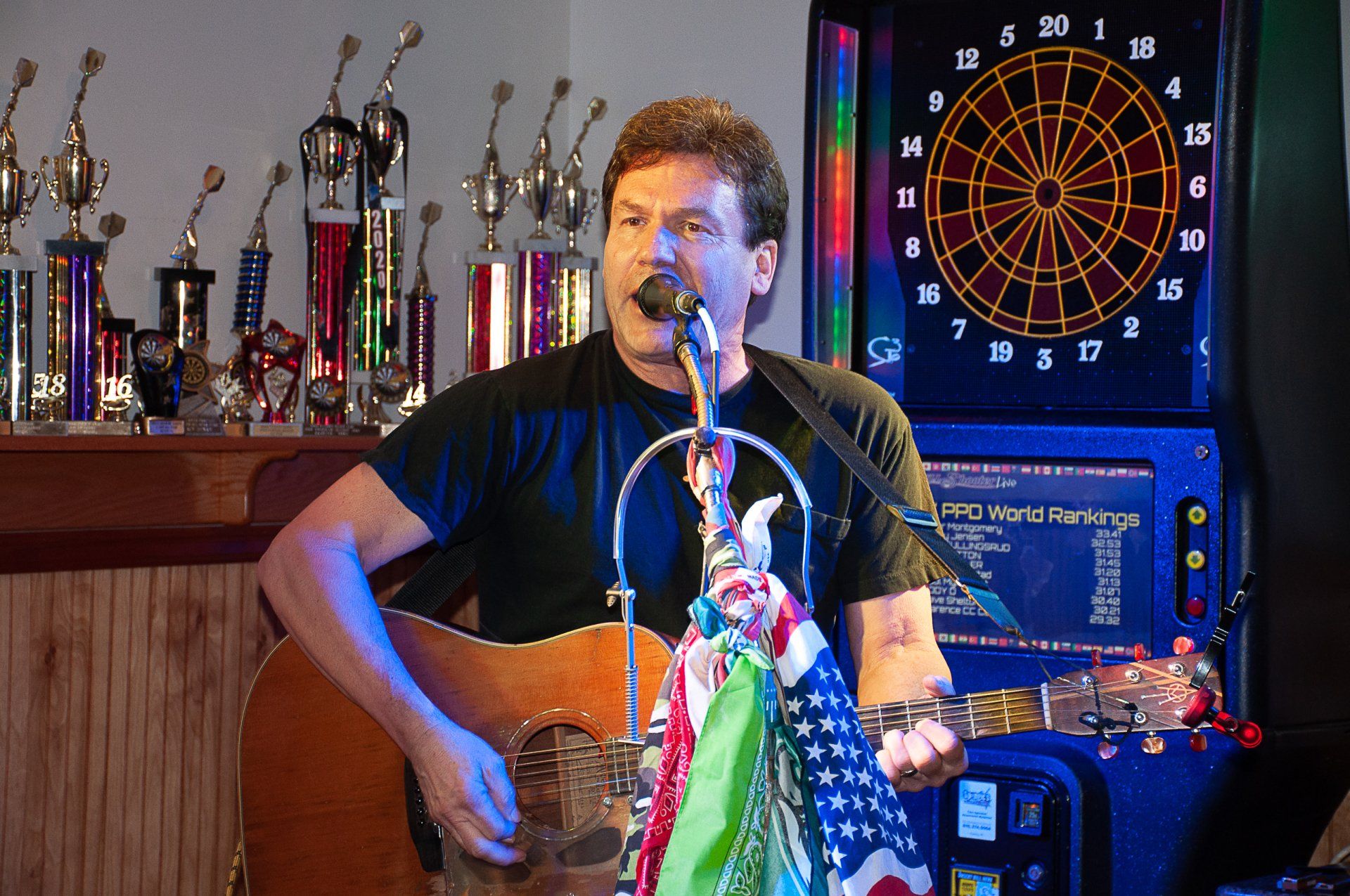 Man playing acoustic guitar and singing onstage, with trophies and a dartboard in the background