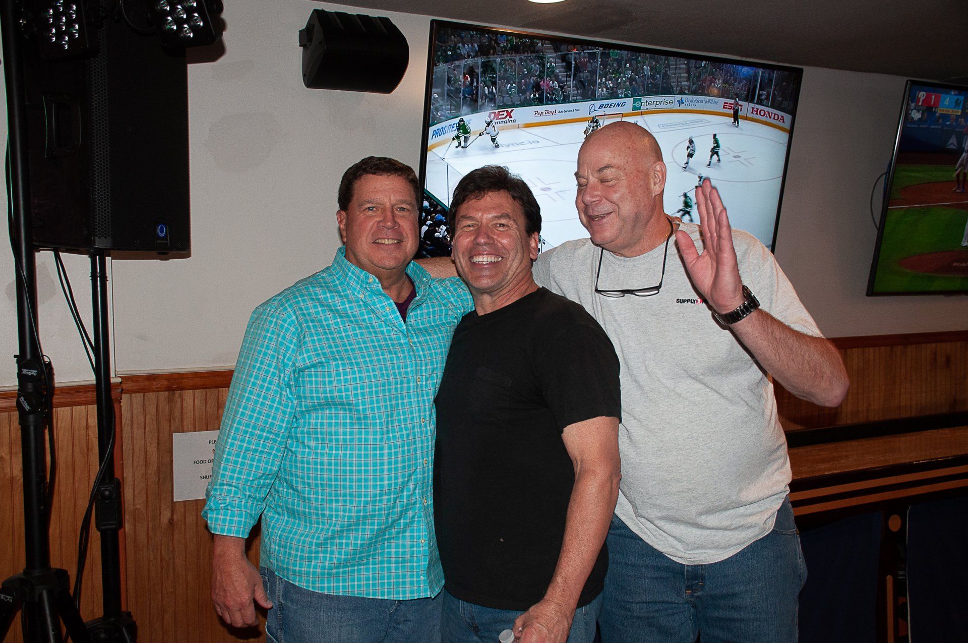 Three men posing in a bar, one waving, with a TV showing a hockey game in the background