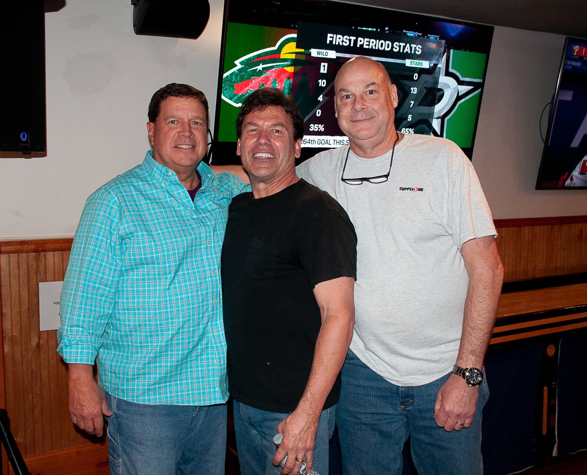 Three men standing together in a bar, smiling in front of a neon sports sign.