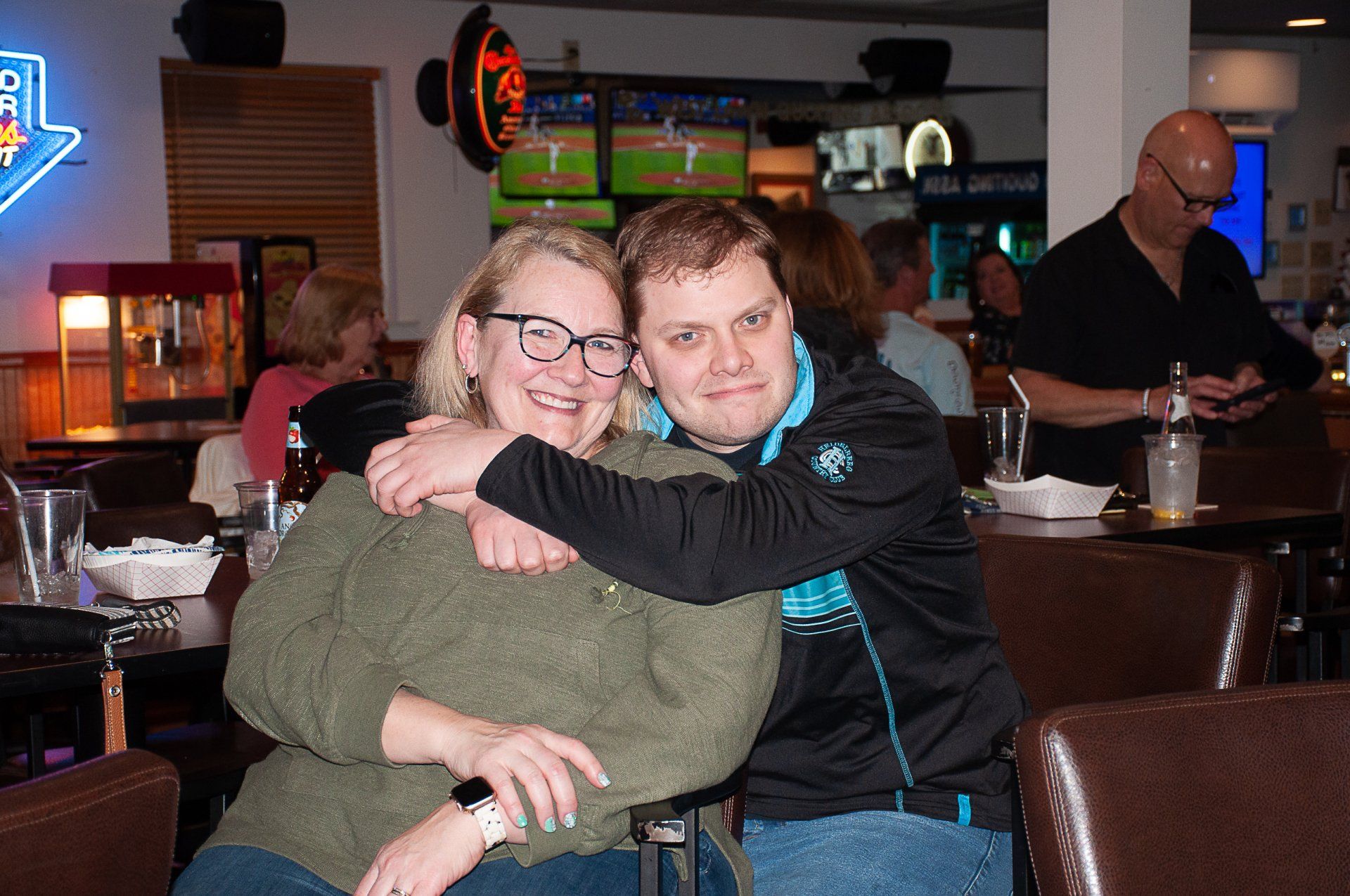 Two people smiling and hugging at a bar with neon lights in the background