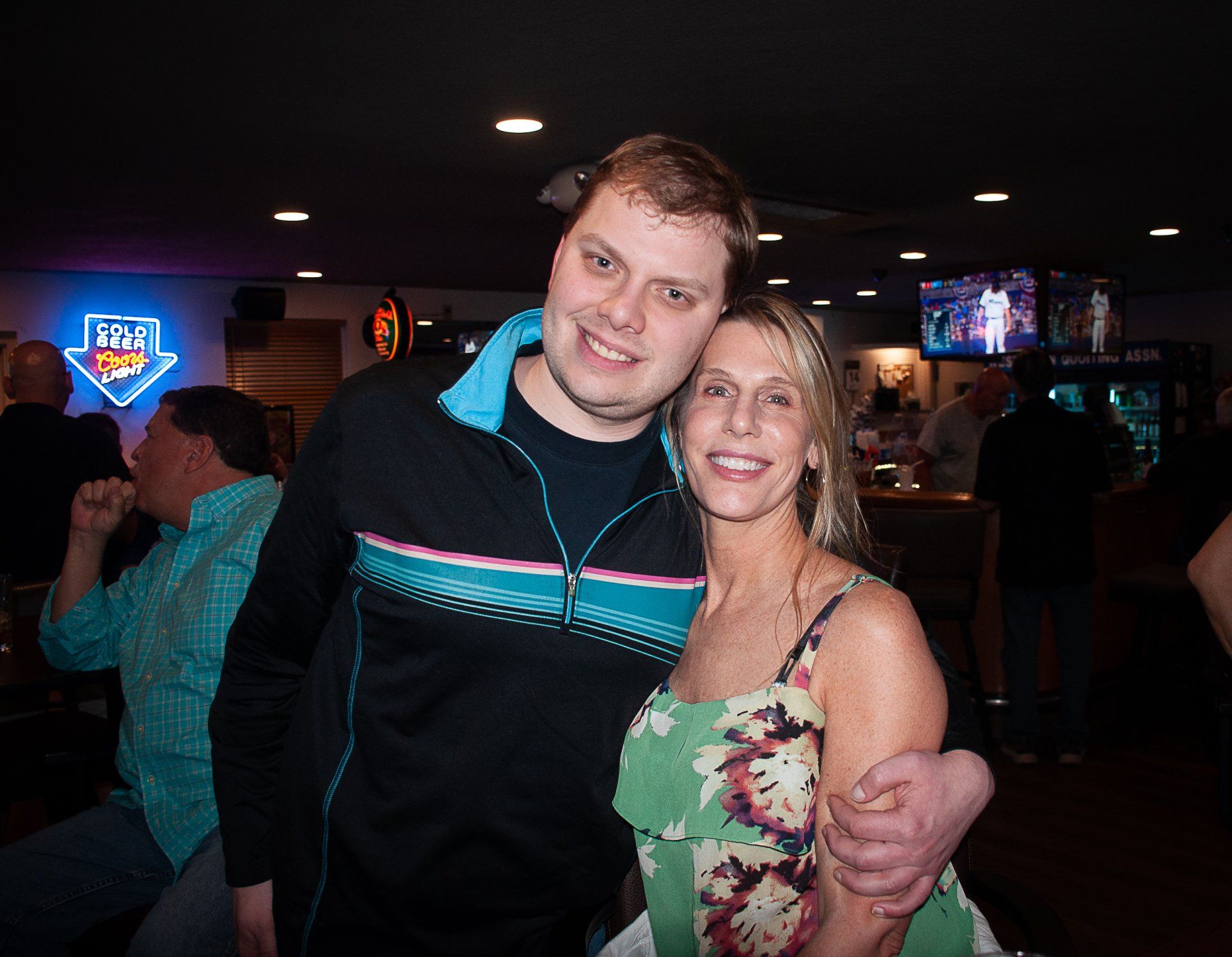 Two people smile and pose together in a dim bar with neon signs in the background.