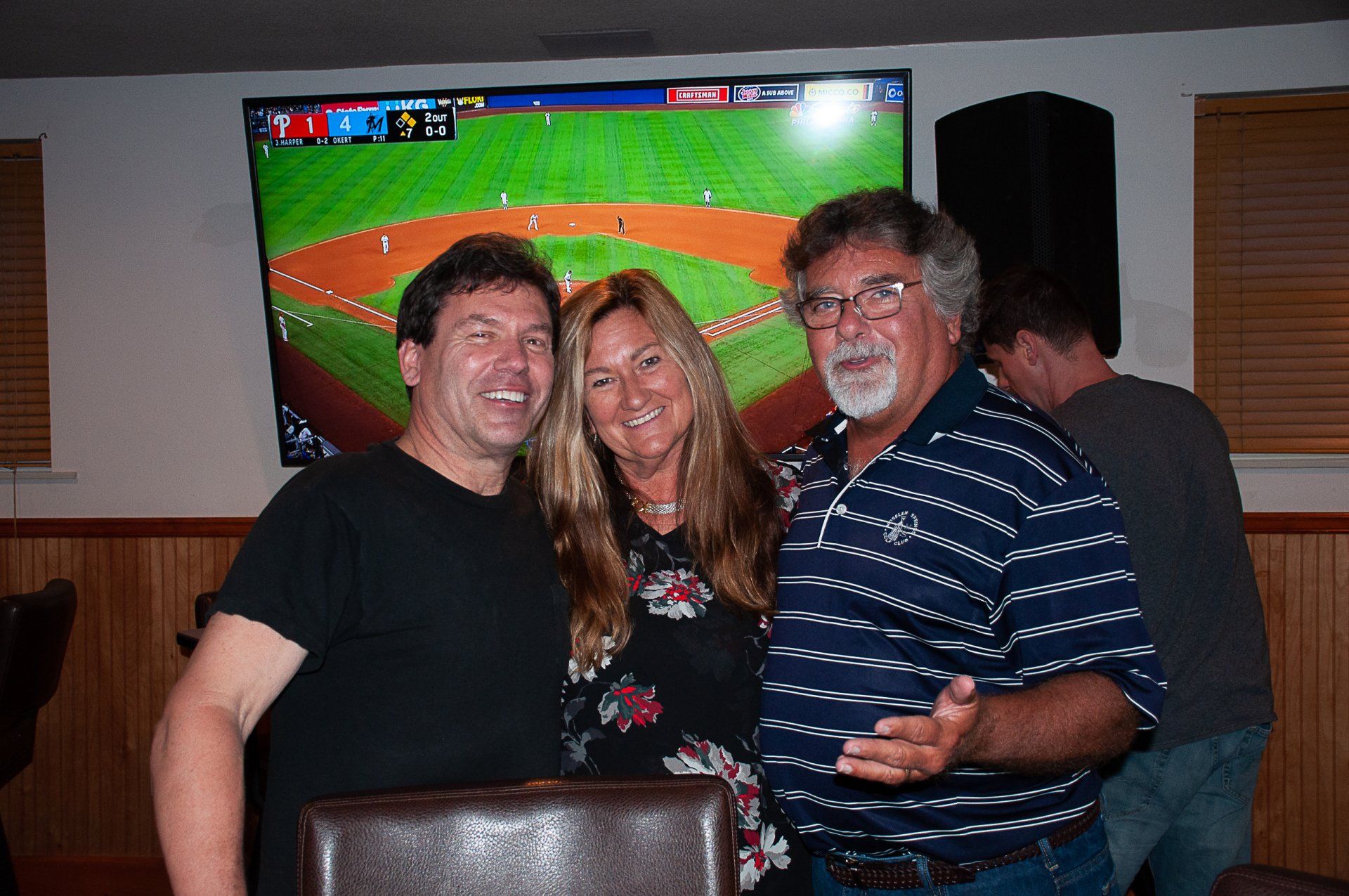 Three smiling people posing indoors in front of a TV showing a baseball game