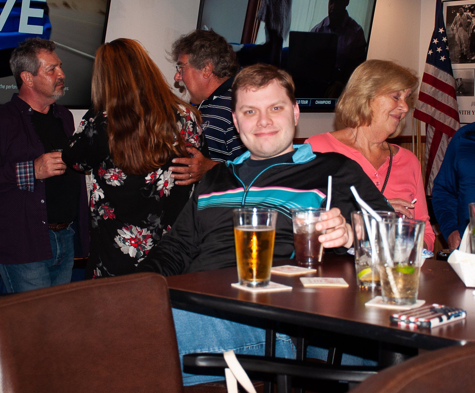 Man seated at a table in a busy bar, with drinks and people chatting in the background.