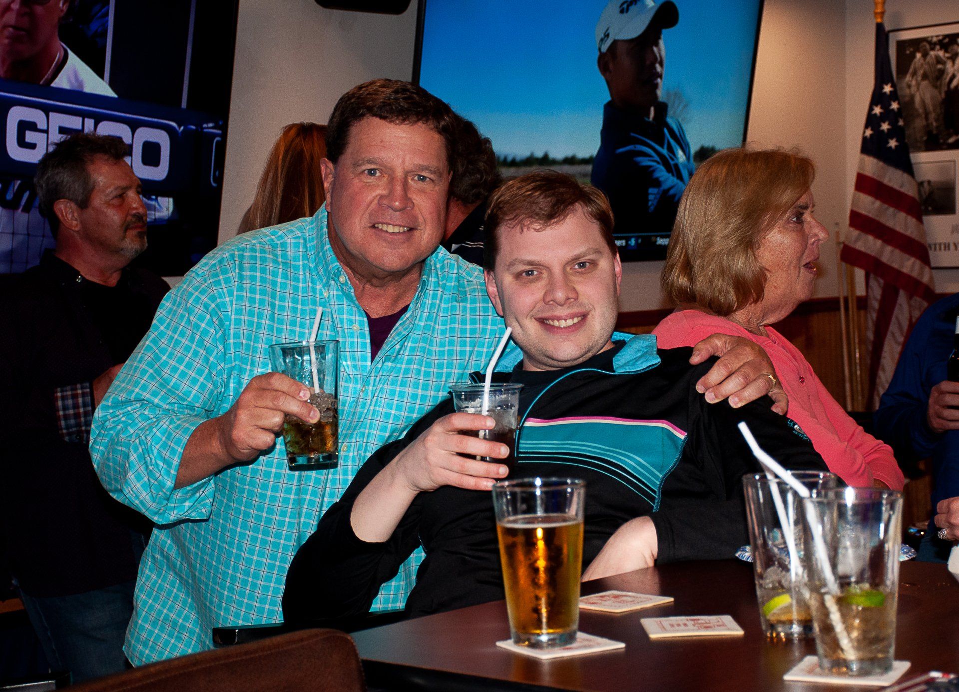 Two people smiling and holding drinks at a bar, with an American flag in the background.