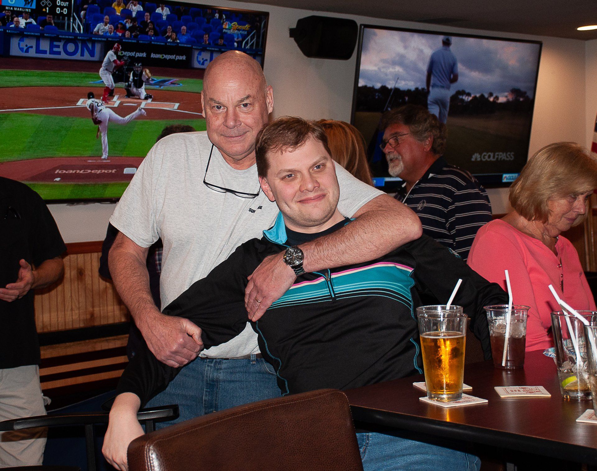 Two men posing at a bar, one hugging the other, with drinks on the table and sports on TV behind them