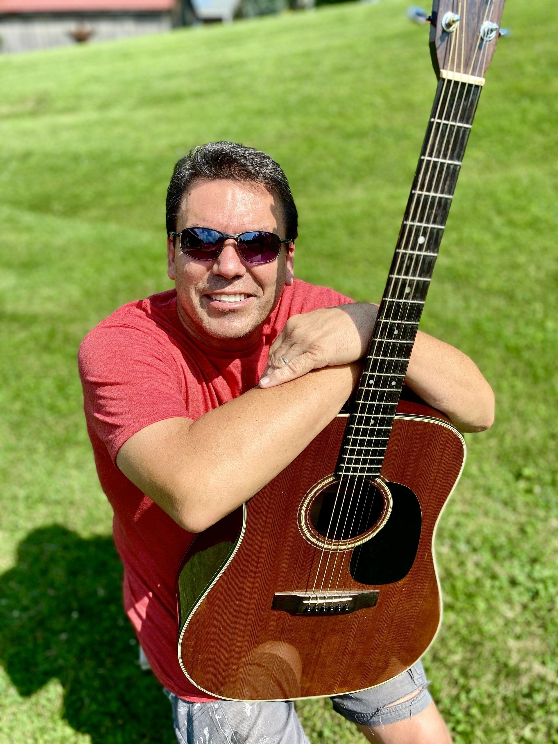 Man wearing sunglasses and a red shirt holding an acoustic guitar on a grassy lawn