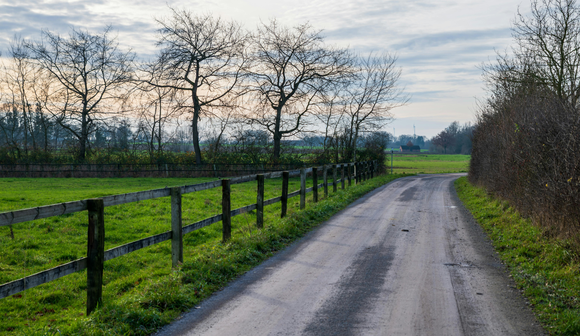 Country road curving past green fields and a black fence under bare trees and a cloudy sky