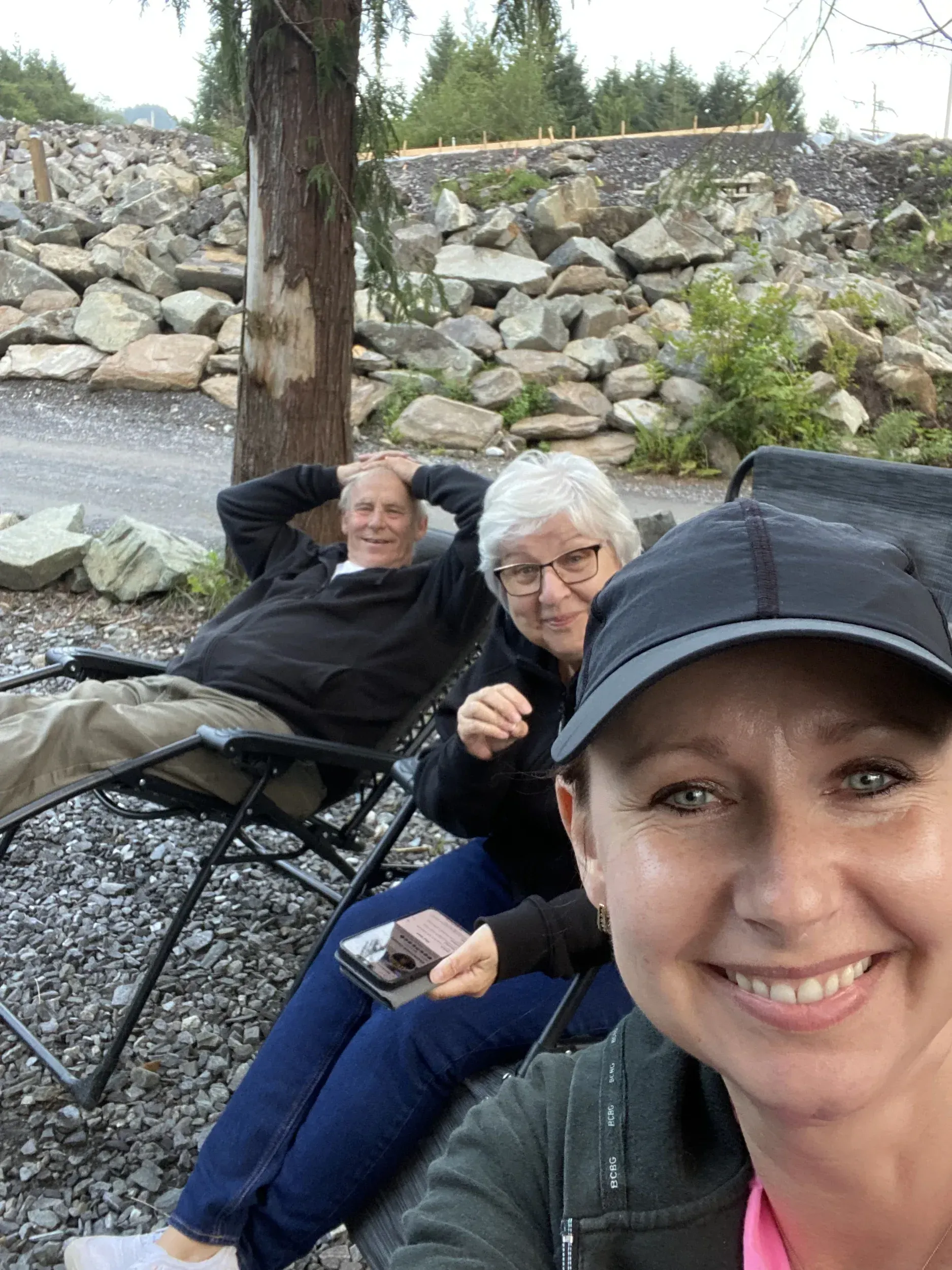 Three people smiling outdoors in folding chairs near rocks and trees, with one taking a selfie