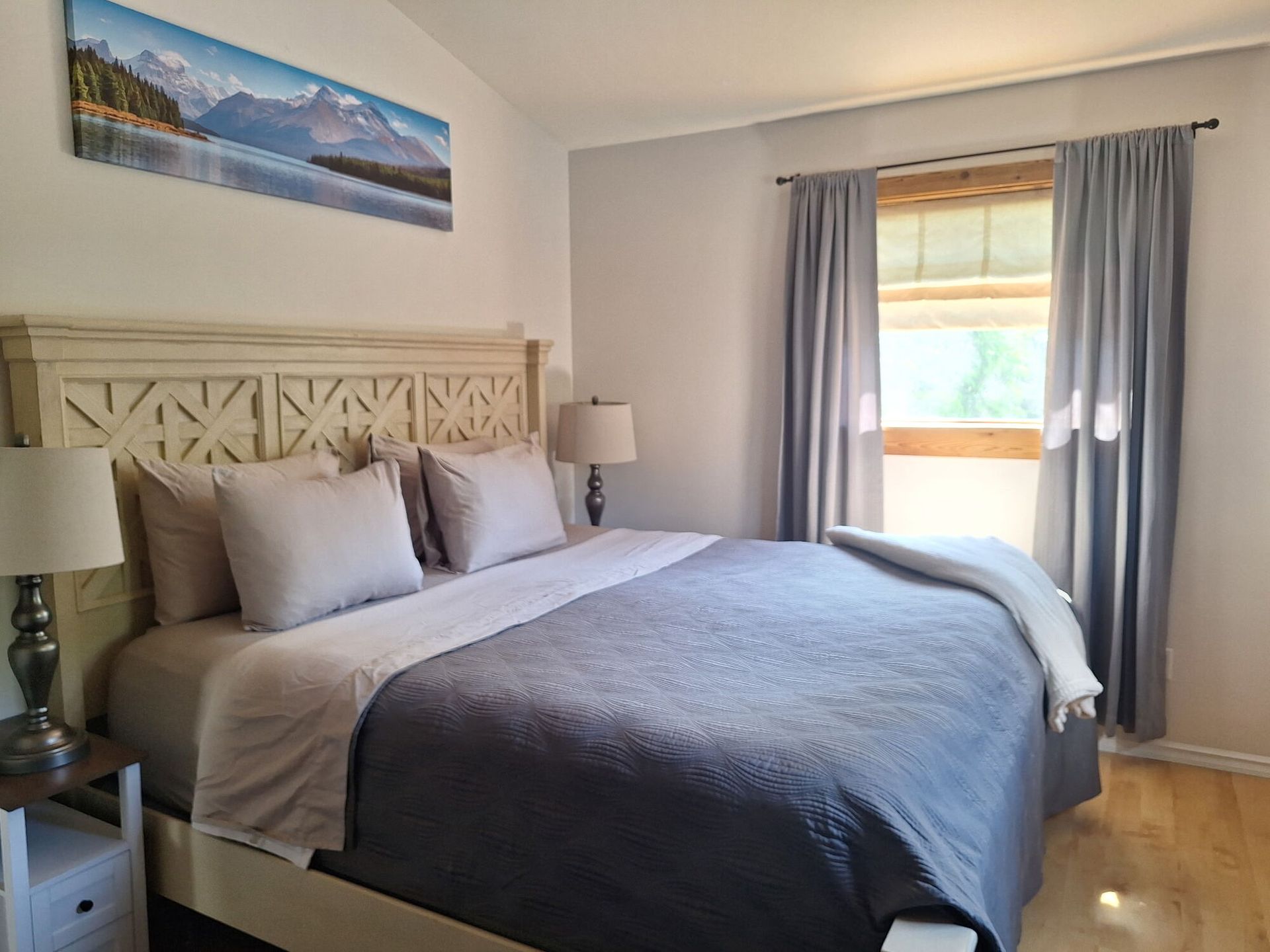 Neatly made bedroom with gray bedding, carved headboard, bedside lamps, and a window with curtains