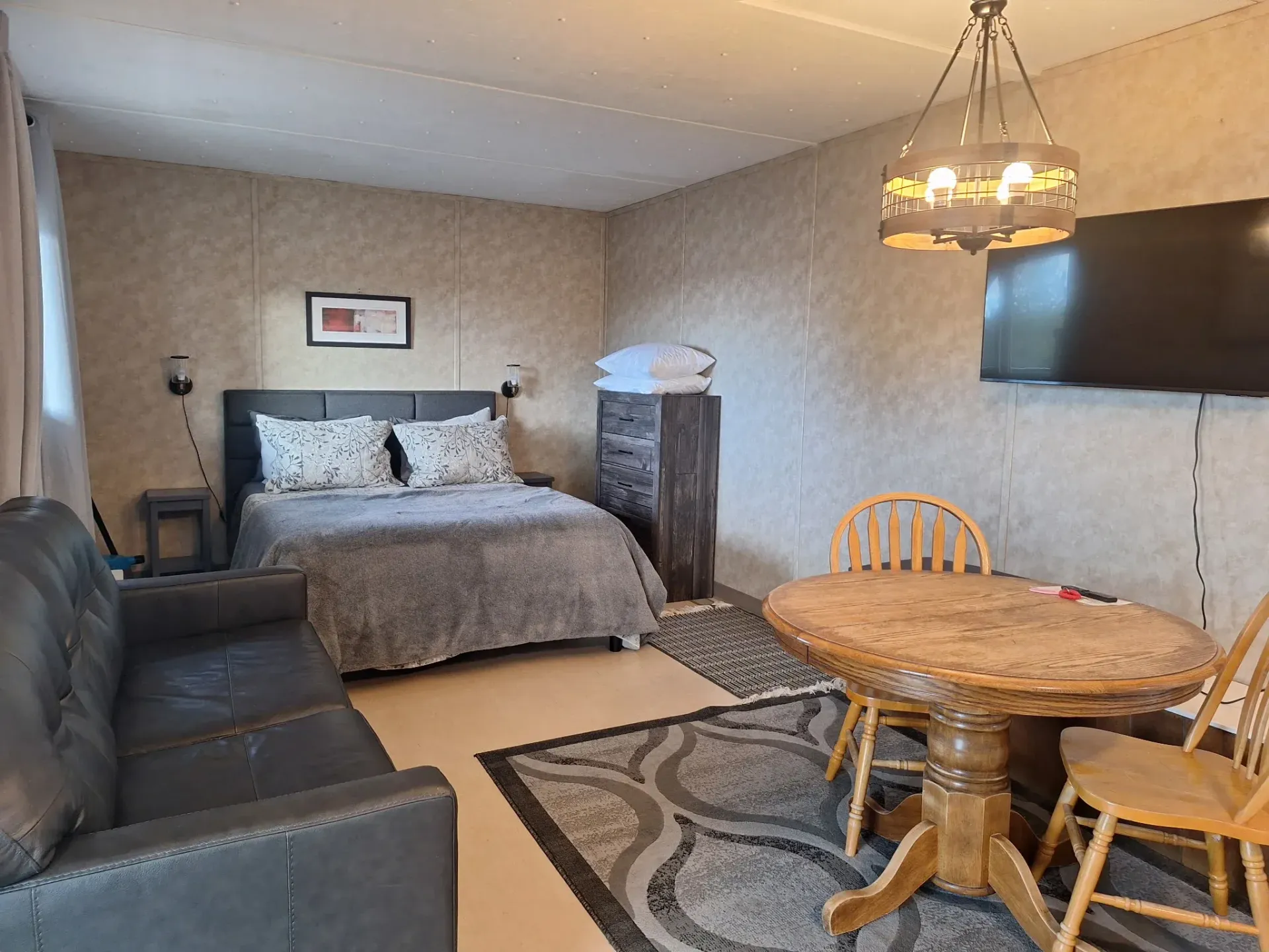Small studio room with bed, sofa, round table, wall TV, and hanging light fixtures