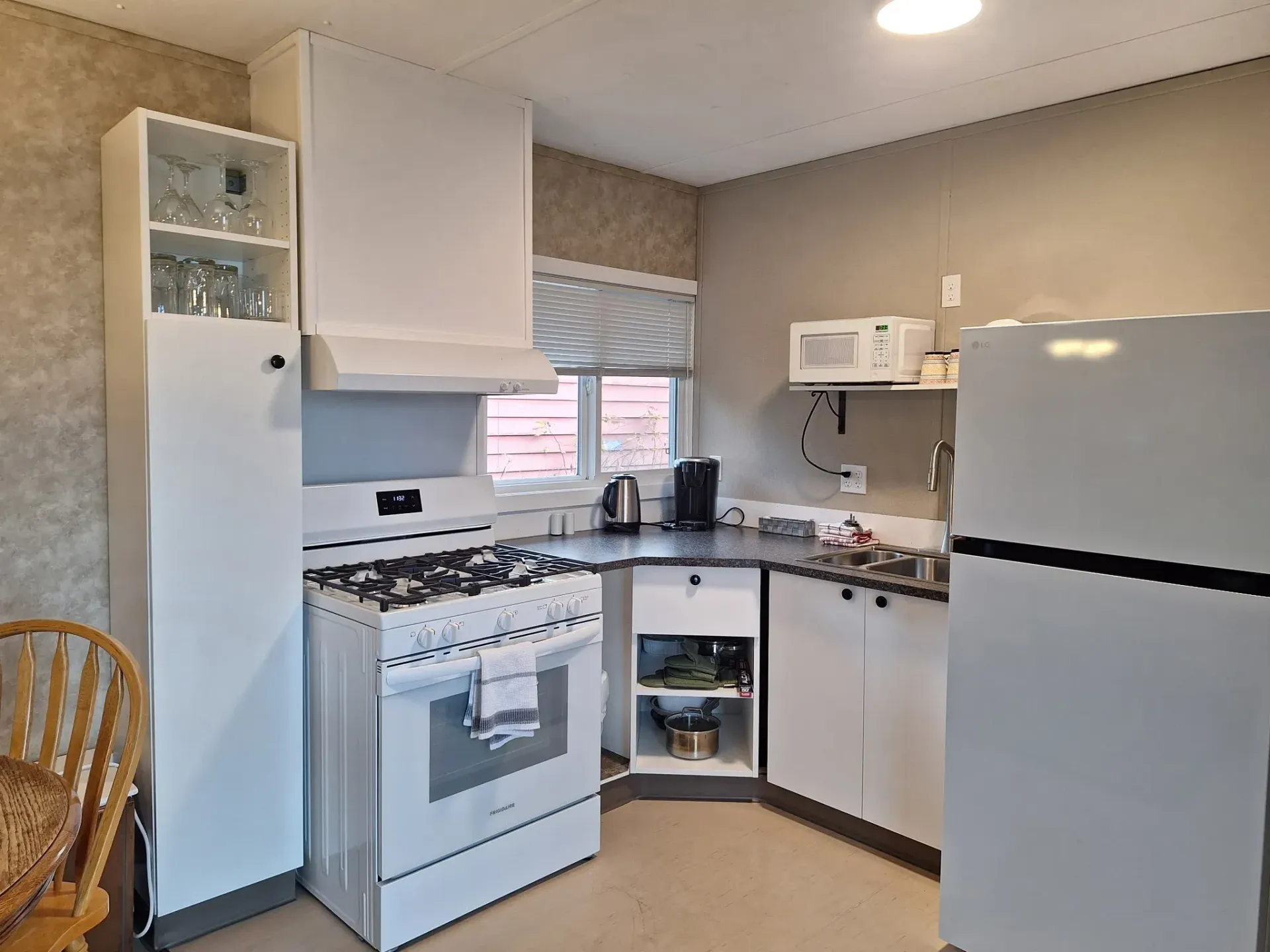 Small white kitchen with stove, fridge, microwave, sink, and corner counter under a window.