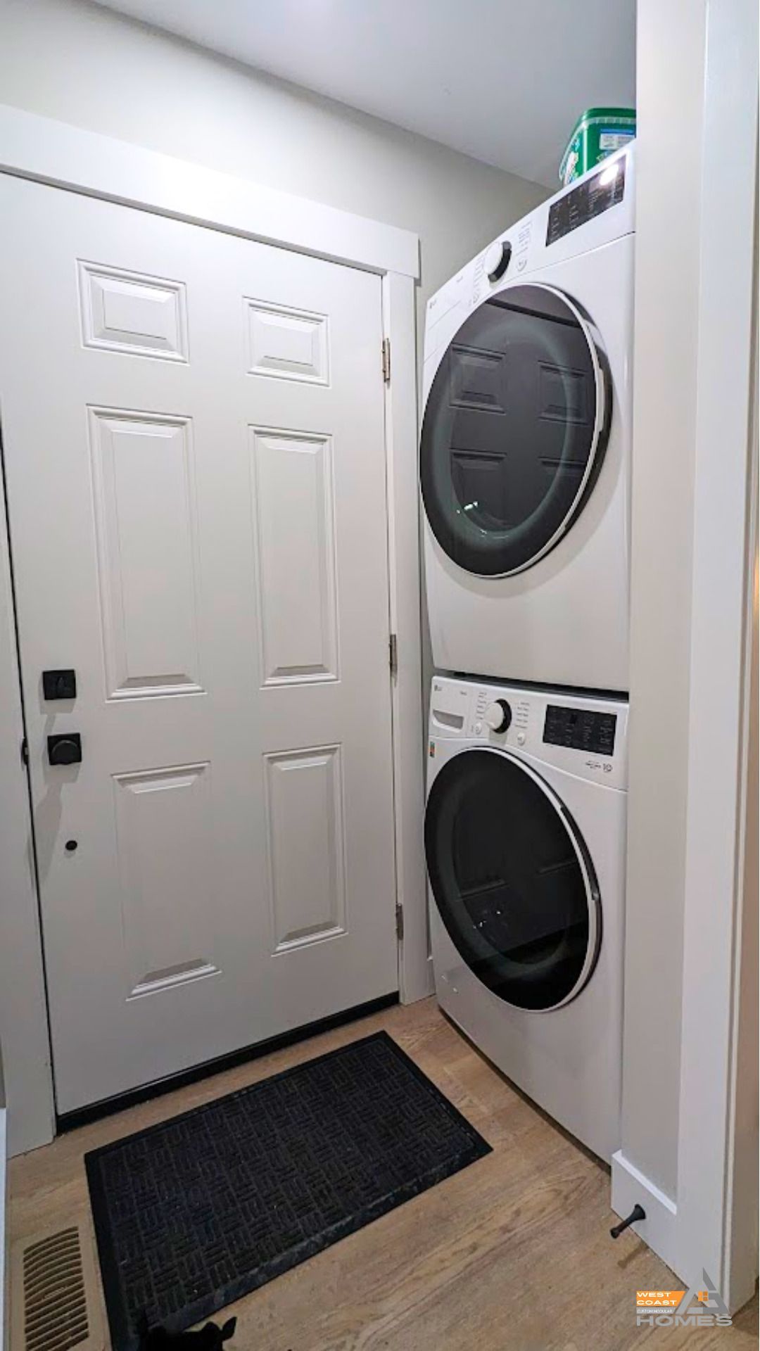 Small laundry area with stacked washer and dryer beside a front door and floor mats.