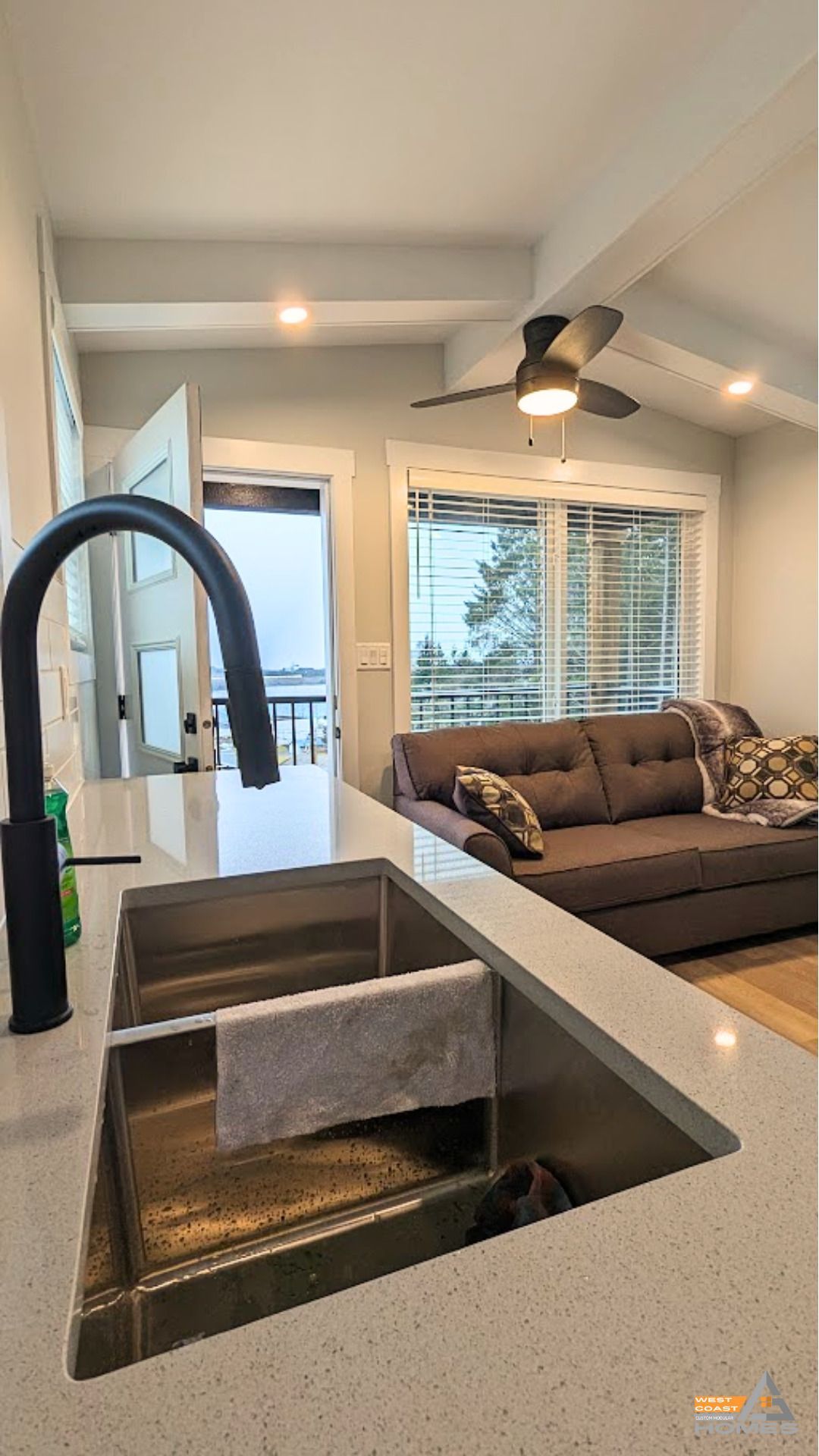 Modern kitchen with farmhouse sink and open living room beyond, featuring a brown sofa and ceiling fan.