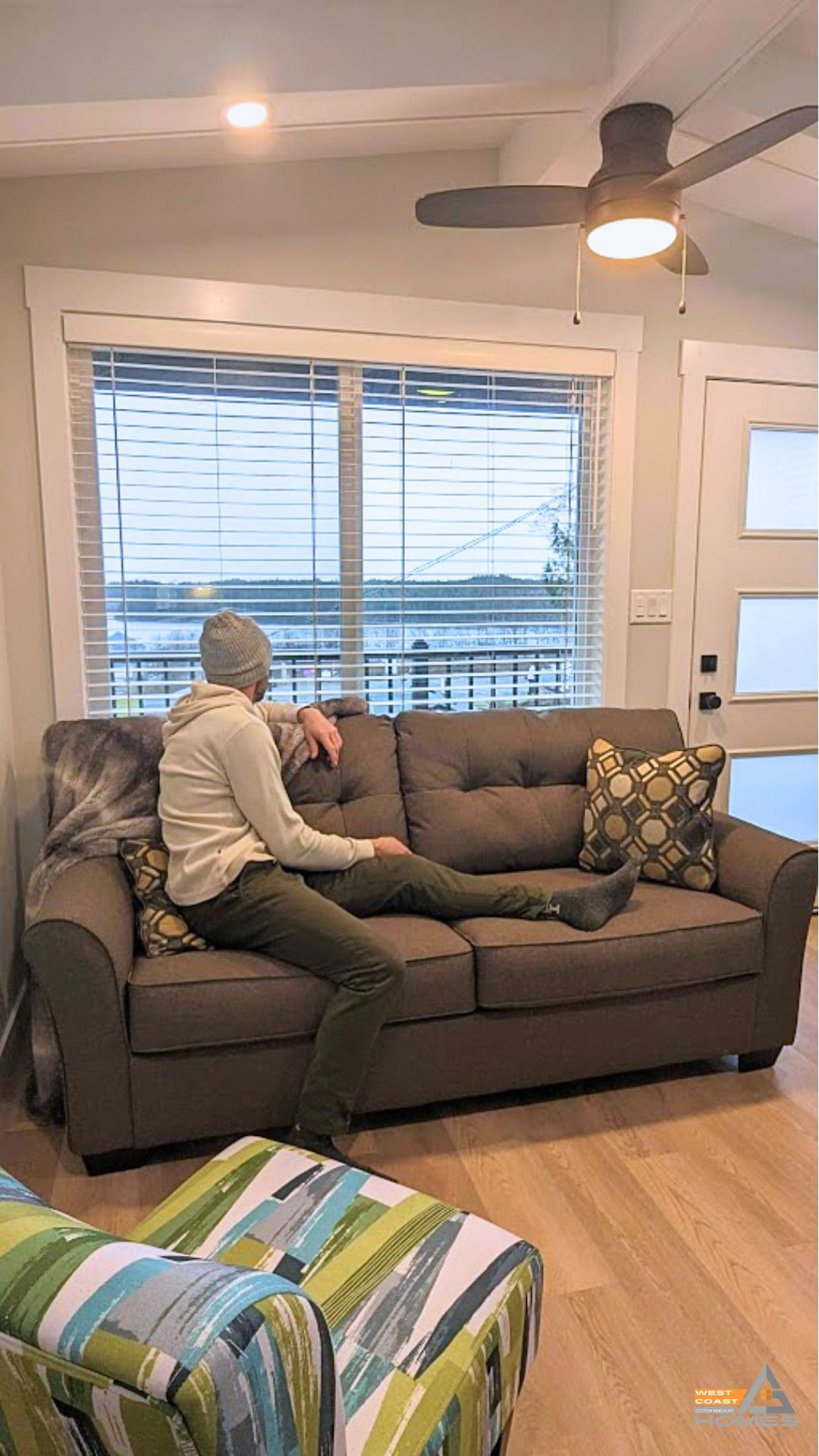 Person sitting on a gray sofa in a bright living room with large windows and a ceiling fan.