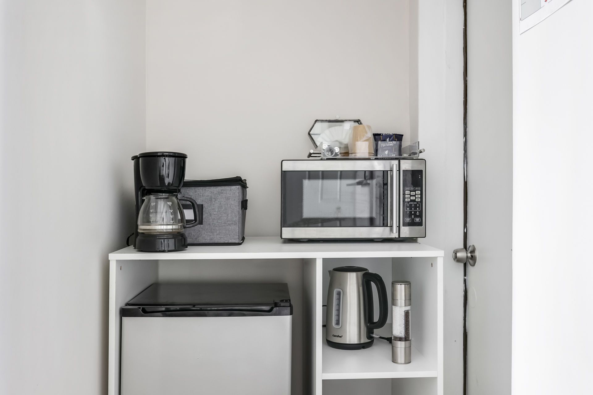 A small kitchenette with a microwave, coffeemaker, mini fridge, and electric kettle on a white shelf.