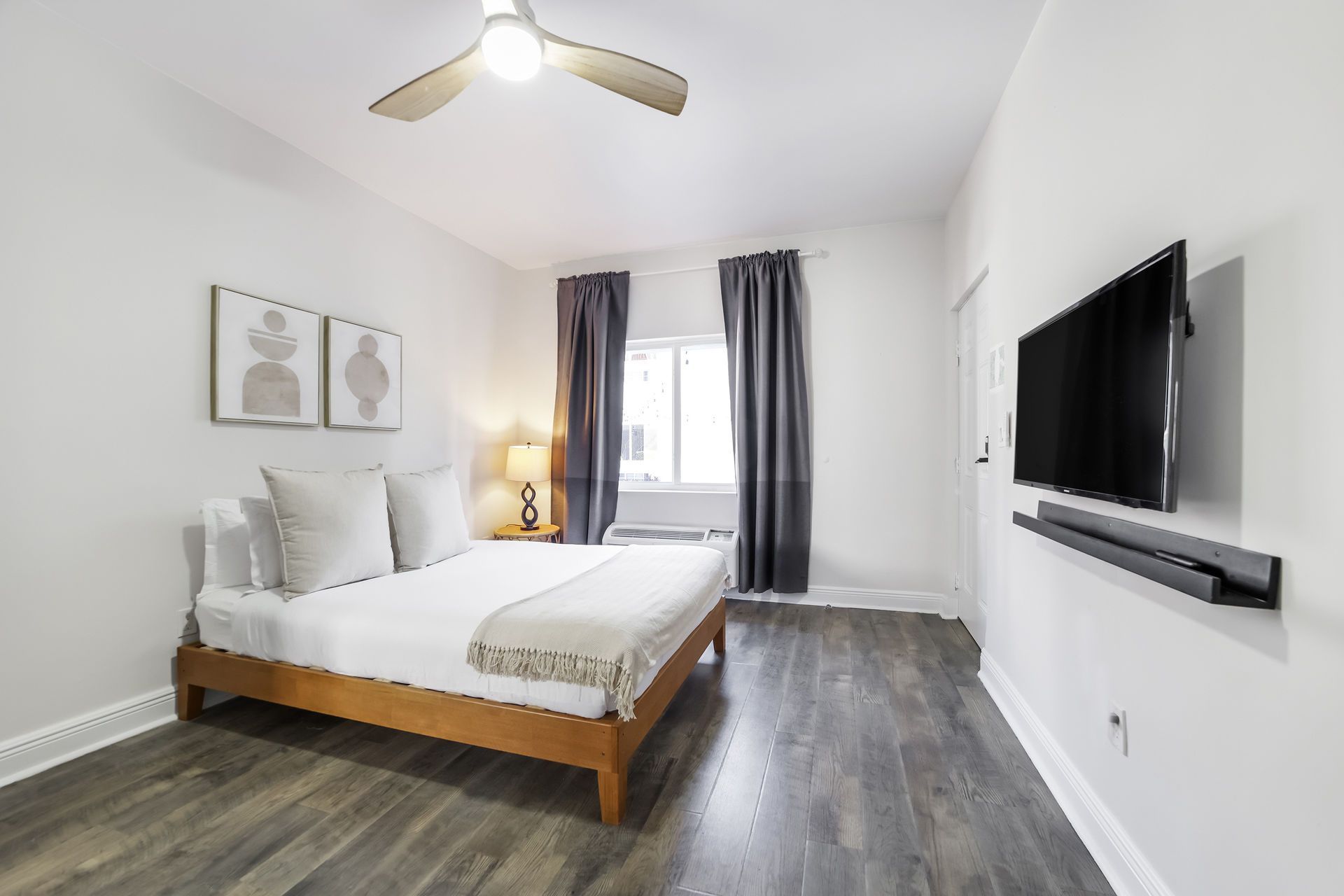 Bedroom with bed, wall-mounted TV, ceiling fan, and window with curtains; light walls, wooden floor.