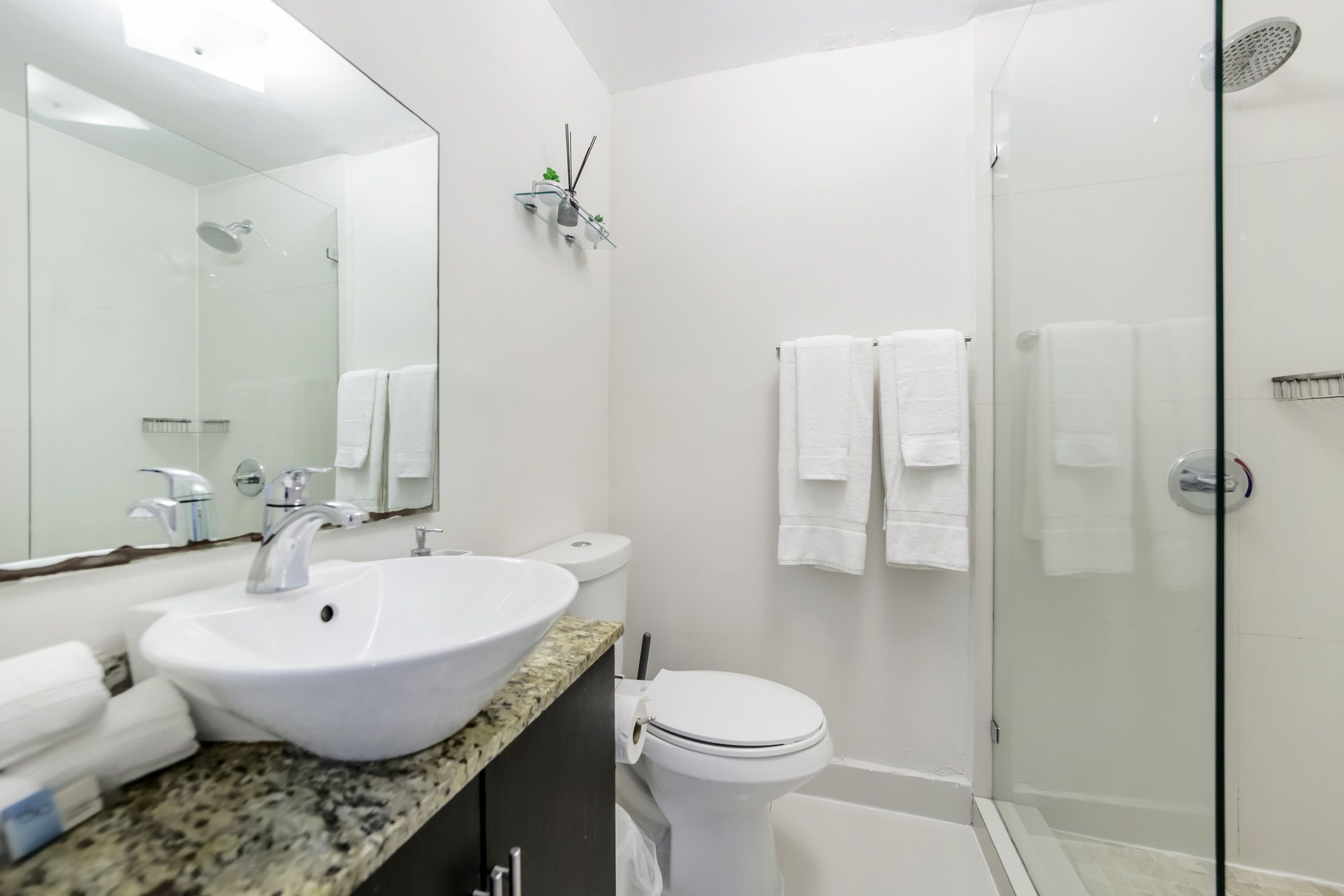 Bathroom with a white toilet, sink, shower, and towels. Gray countertop, mirror, and glass shower door.