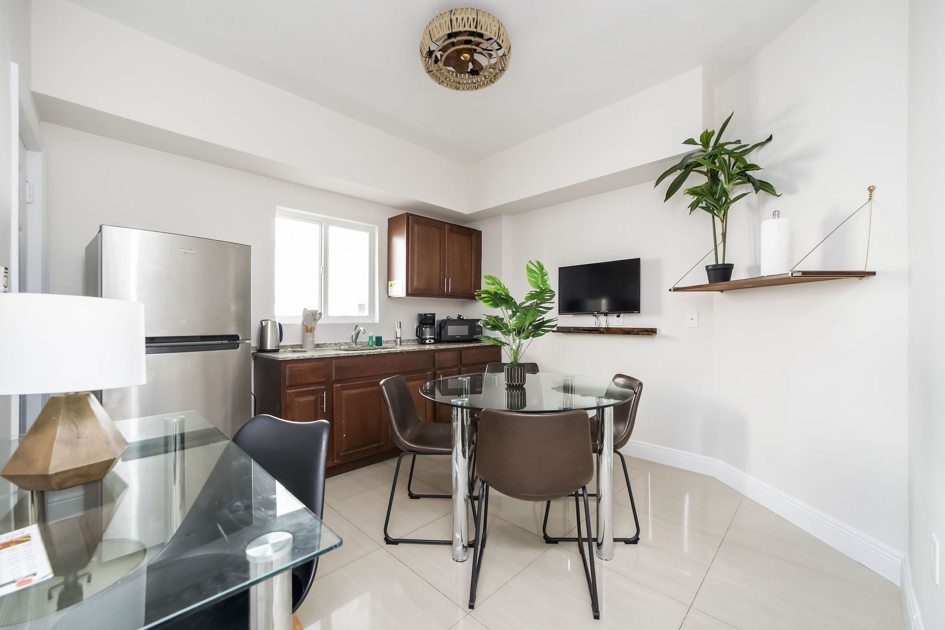 A small kitchen and dining area with stainless steel fridge, dark cabinets, a round table, and a wall-mounted TV.