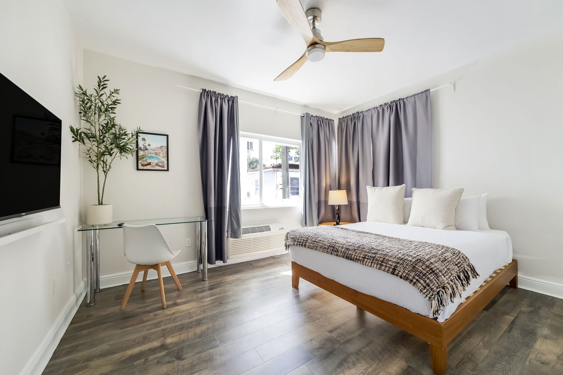 Bedroom with bed, desk, curtains, TV, and ceiling fan. Light wood floors and furniture.