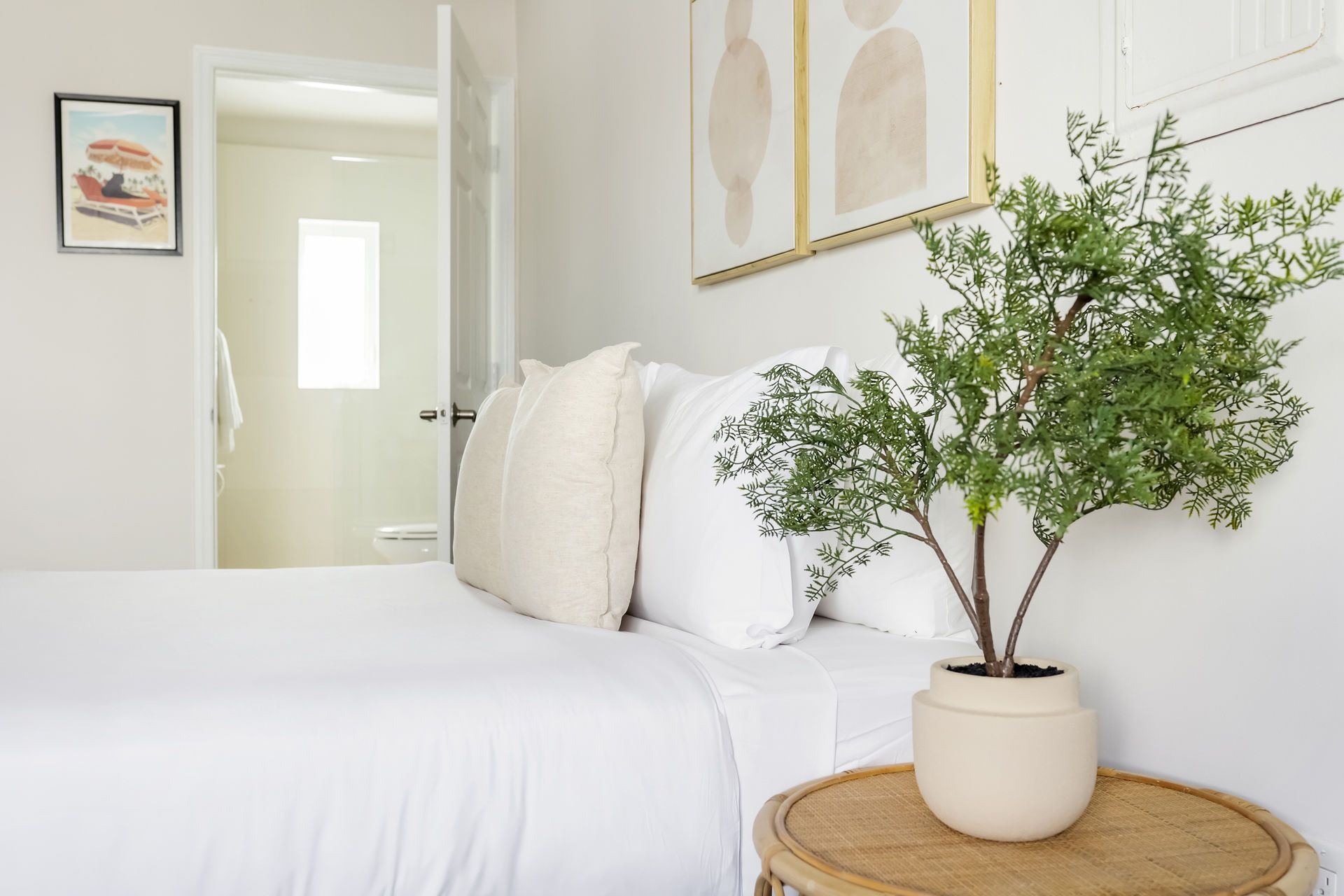 Bedroom with white bed, neutral pillows, artwork, and a potted plant on a woven side table.