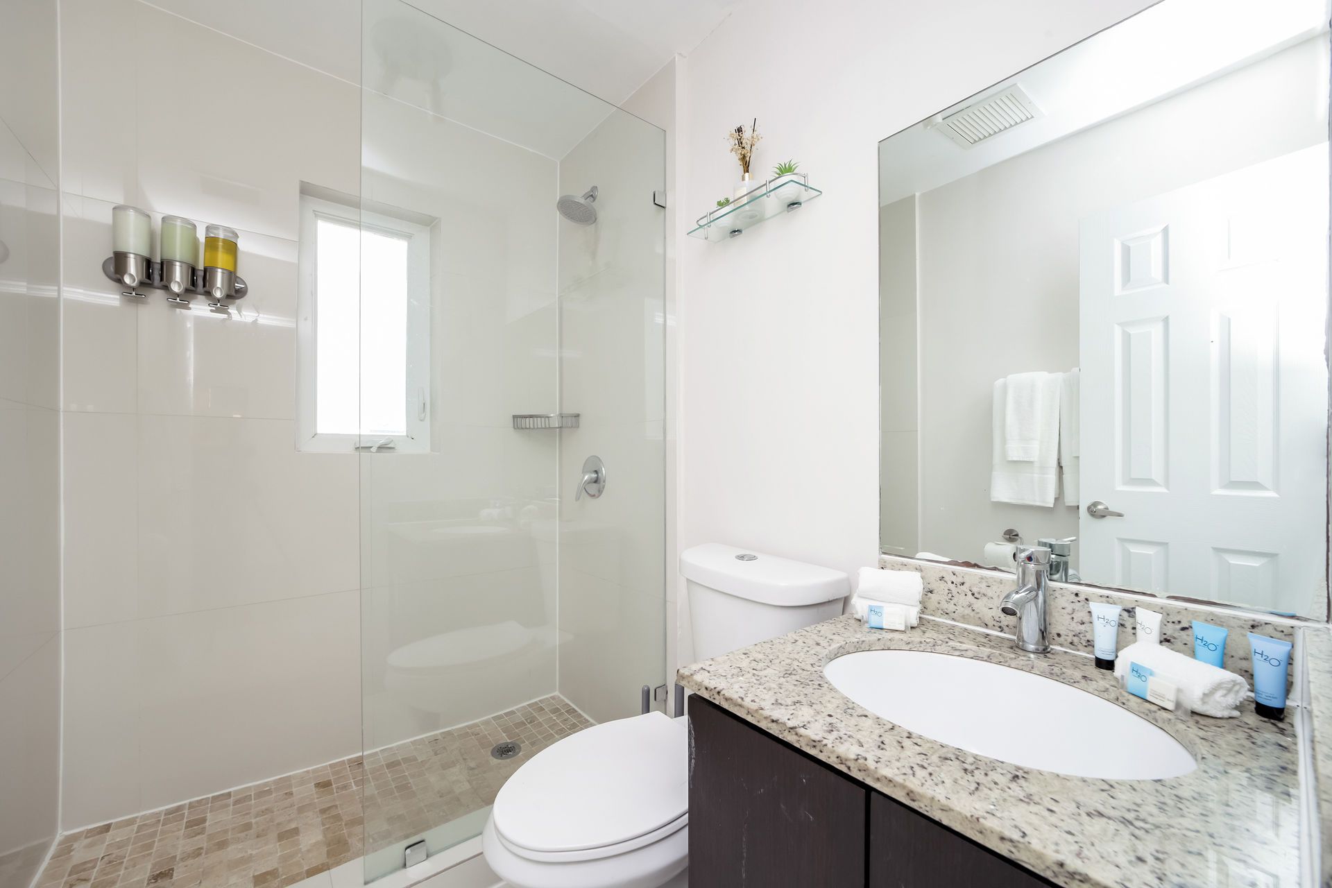 Bathroom with a glass shower, toilet, vanity, and a window. Light colors, towels, and toiletries are visible.