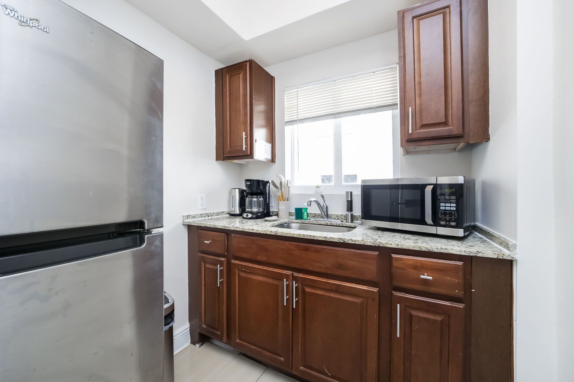 Small kitchen with stainless steel refrigerator, brown cabinets, and a microwave next to a window.