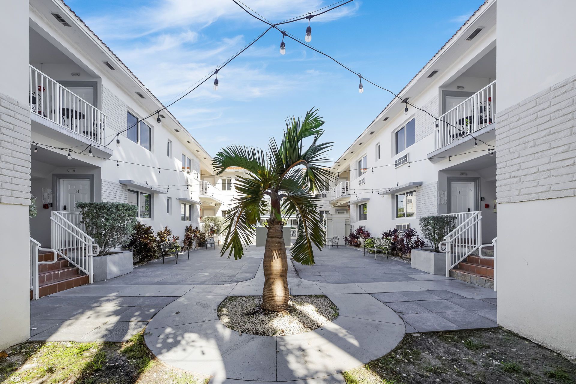 Courtyard with two-story white buildings, a palm tree, and string lights, with a brick walkway and greenery.