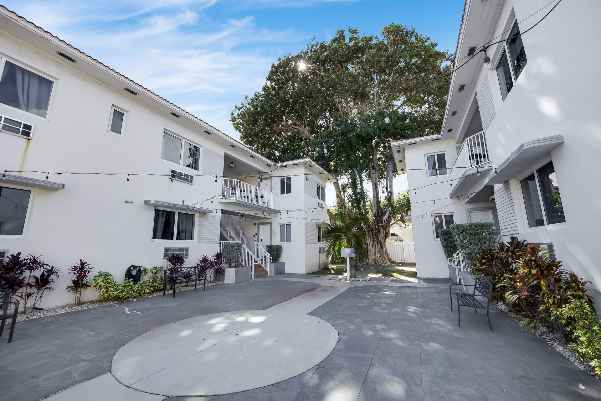 White apartment buildings with gray patio, lush plants, and a large tree under blue sky.