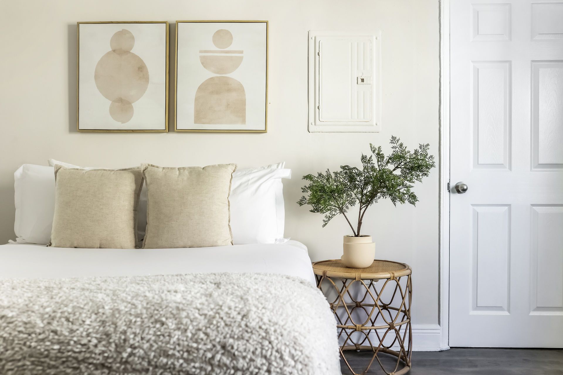 Bedroom with neutral tones. Bed with textured blanket and pillows, artwork, a small table, and a tree. White door.