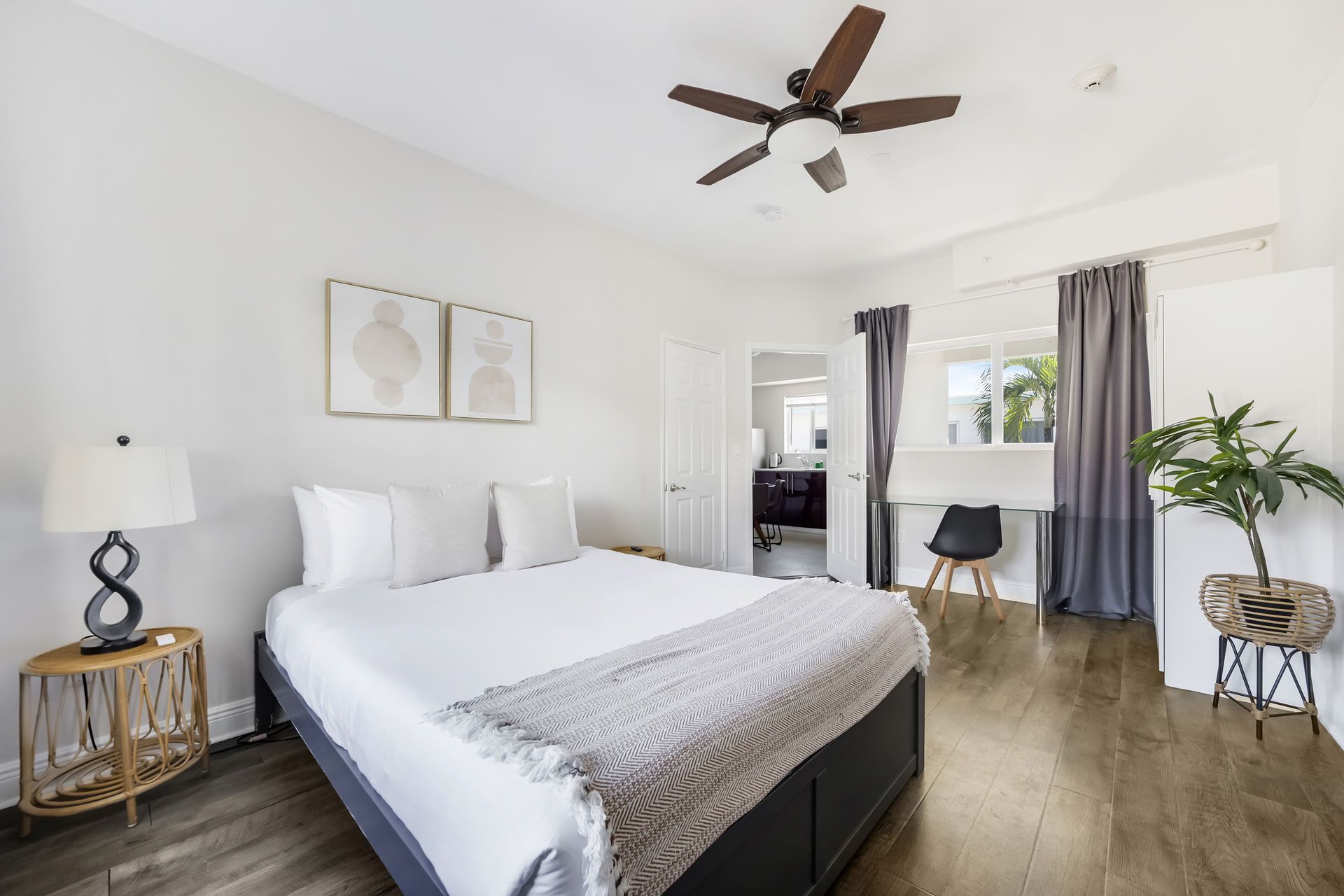 Bedroom with a bed, nightstand, desk, and a potted plant. The room has white walls and a dark ceiling fan.