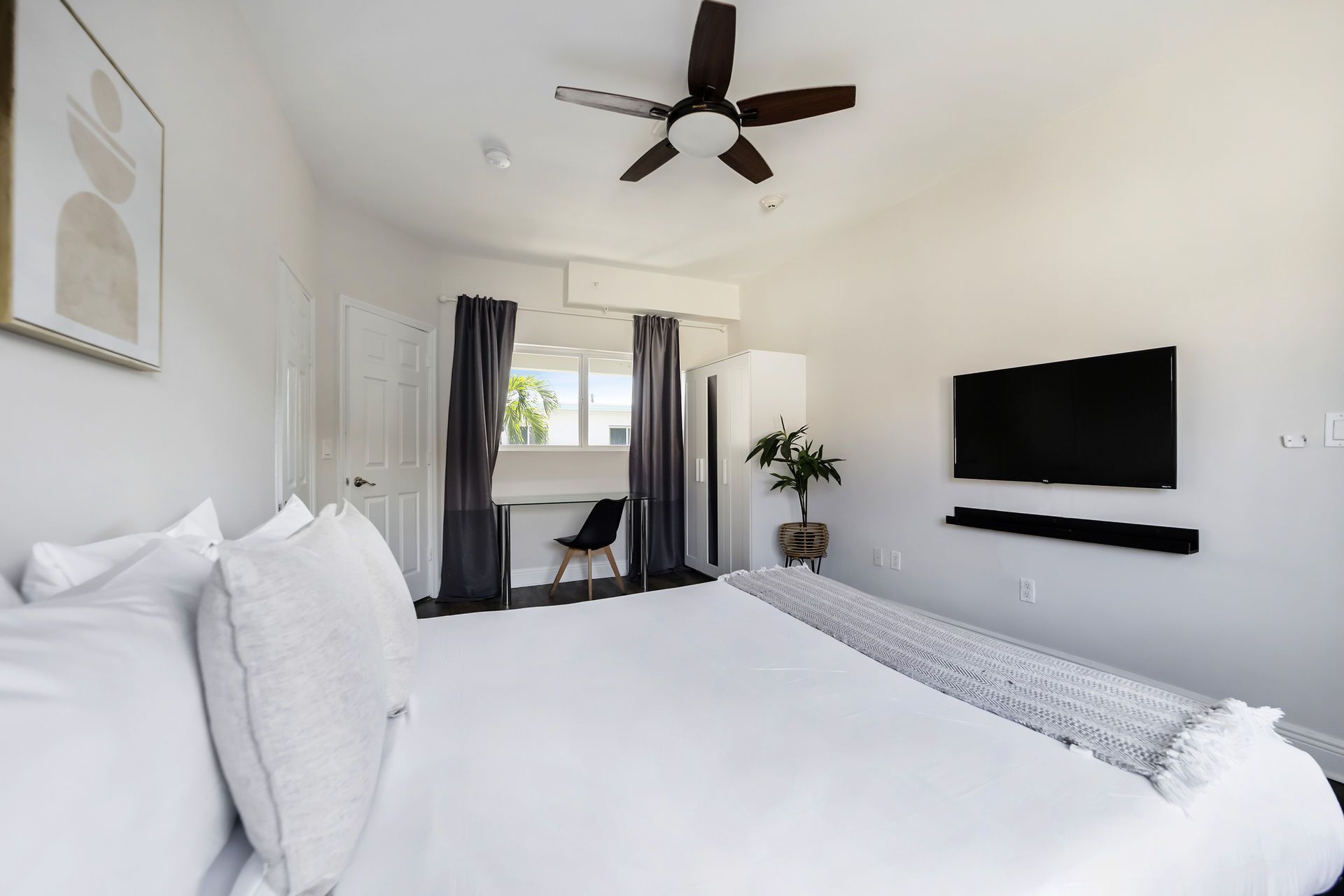 Bedroom with a large bed, TV, desk, and ceiling fan. White walls, gray curtains, and a plant.