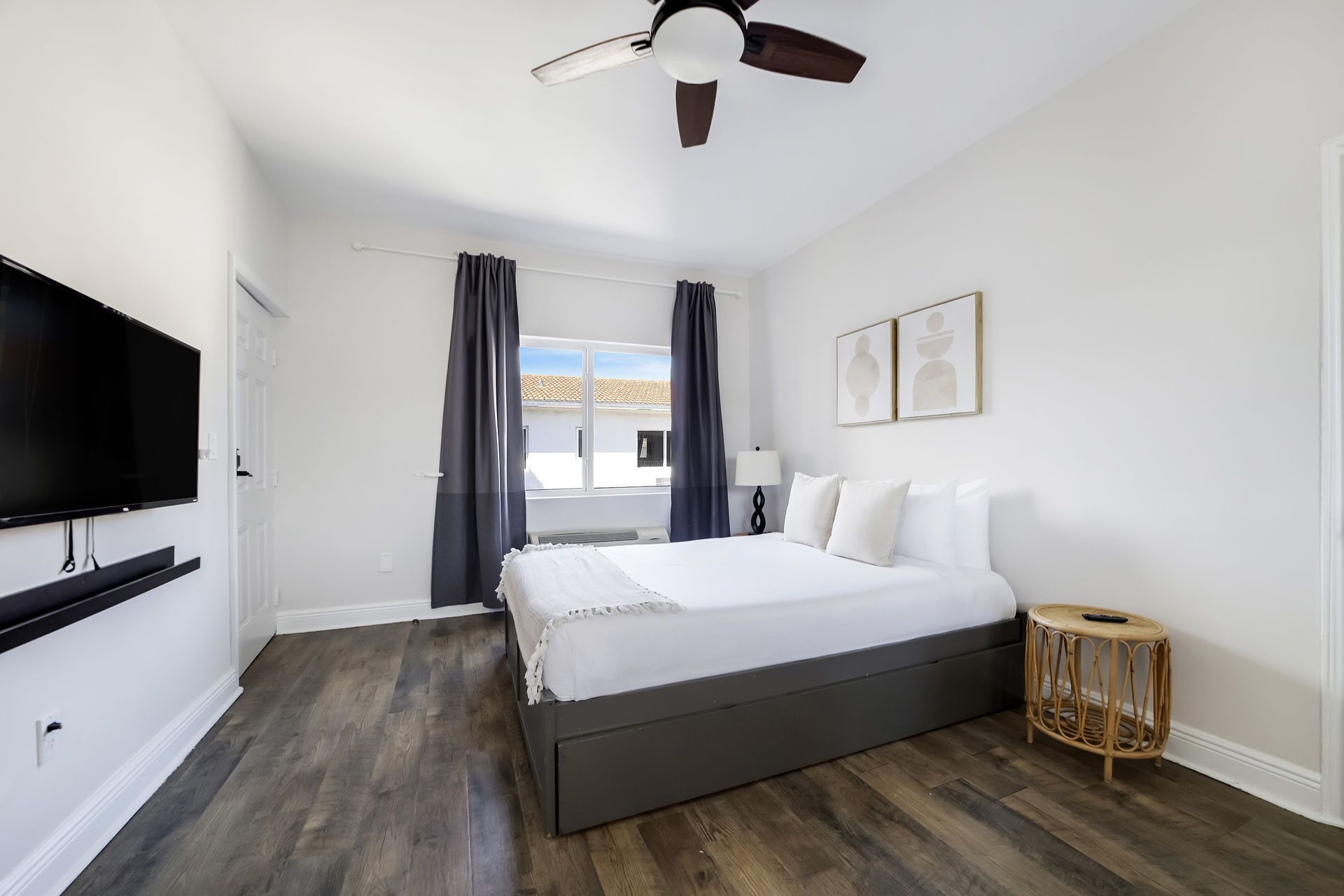 Bedroom with a bed, TV, window, and small side table. Dark flooring, white walls, and neutral decor.