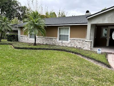 A single-story house with tan stucco, gray roof, stone facade, and palm trees in a green lawn.