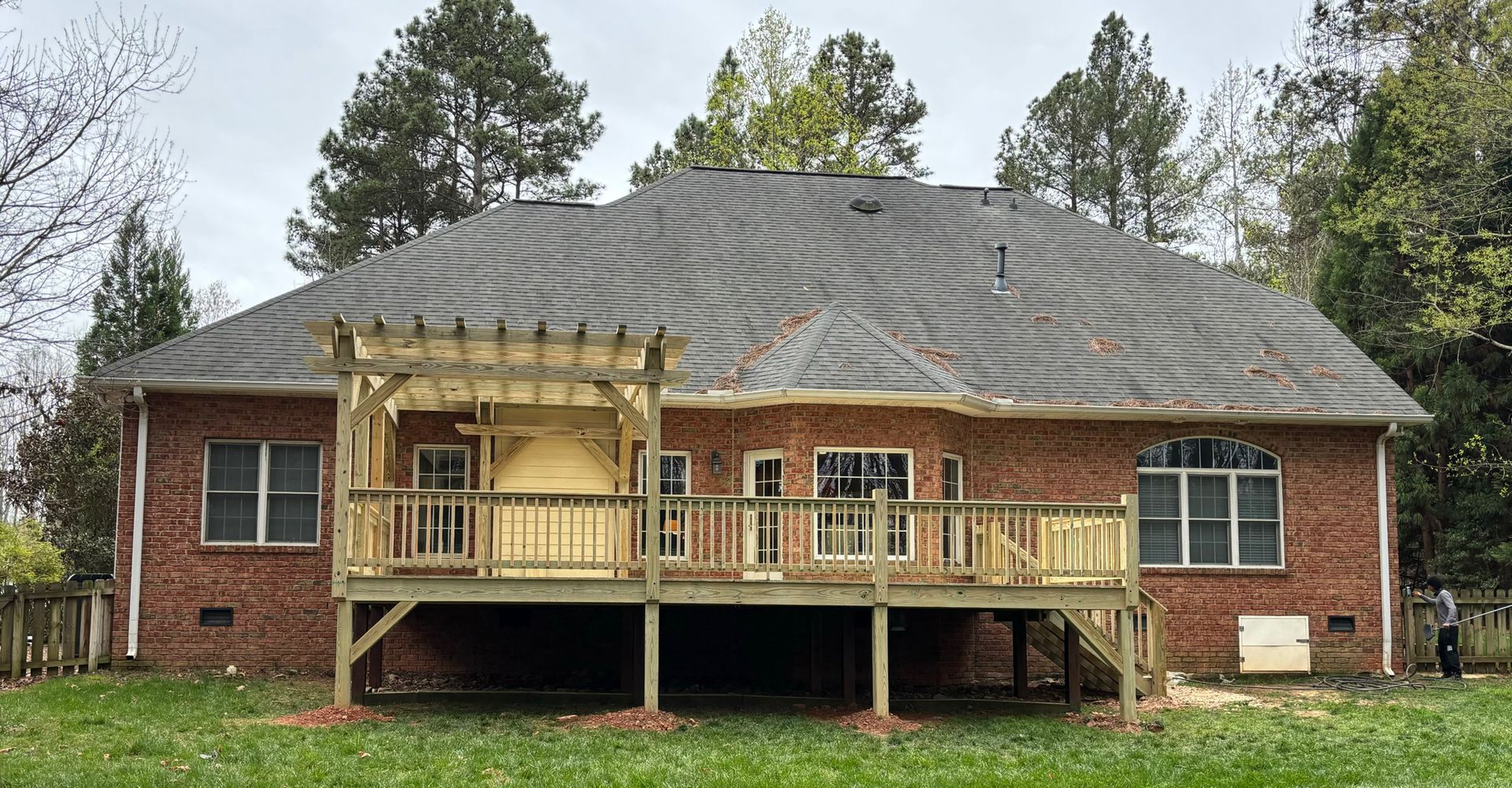 Back of a brick house with a wooden deck and pergola, set on a green lawn with a tree-lined background.
