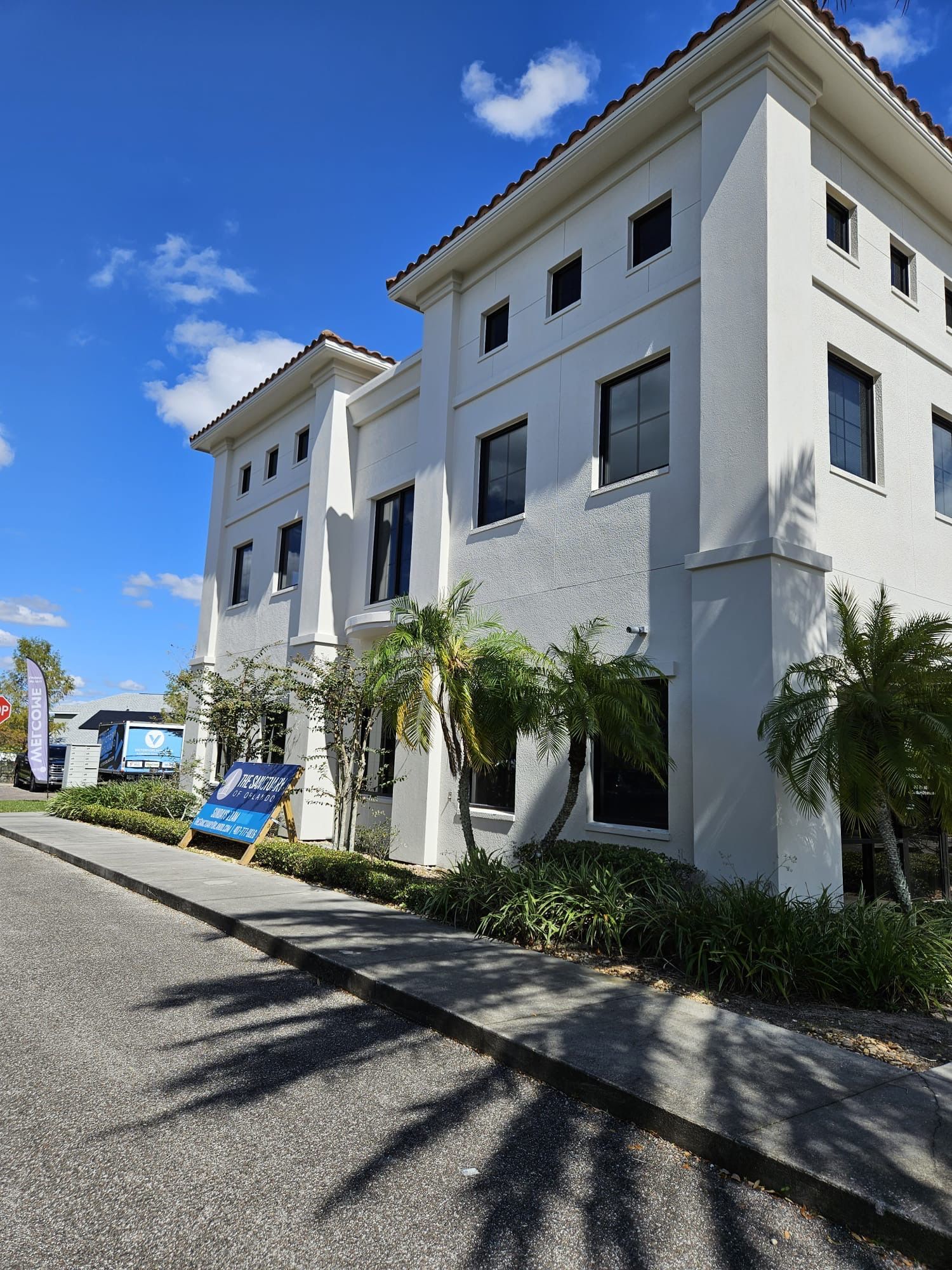 White two-story building with dark windows and a tile roof, palm trees, and a sign on the sidewalk.