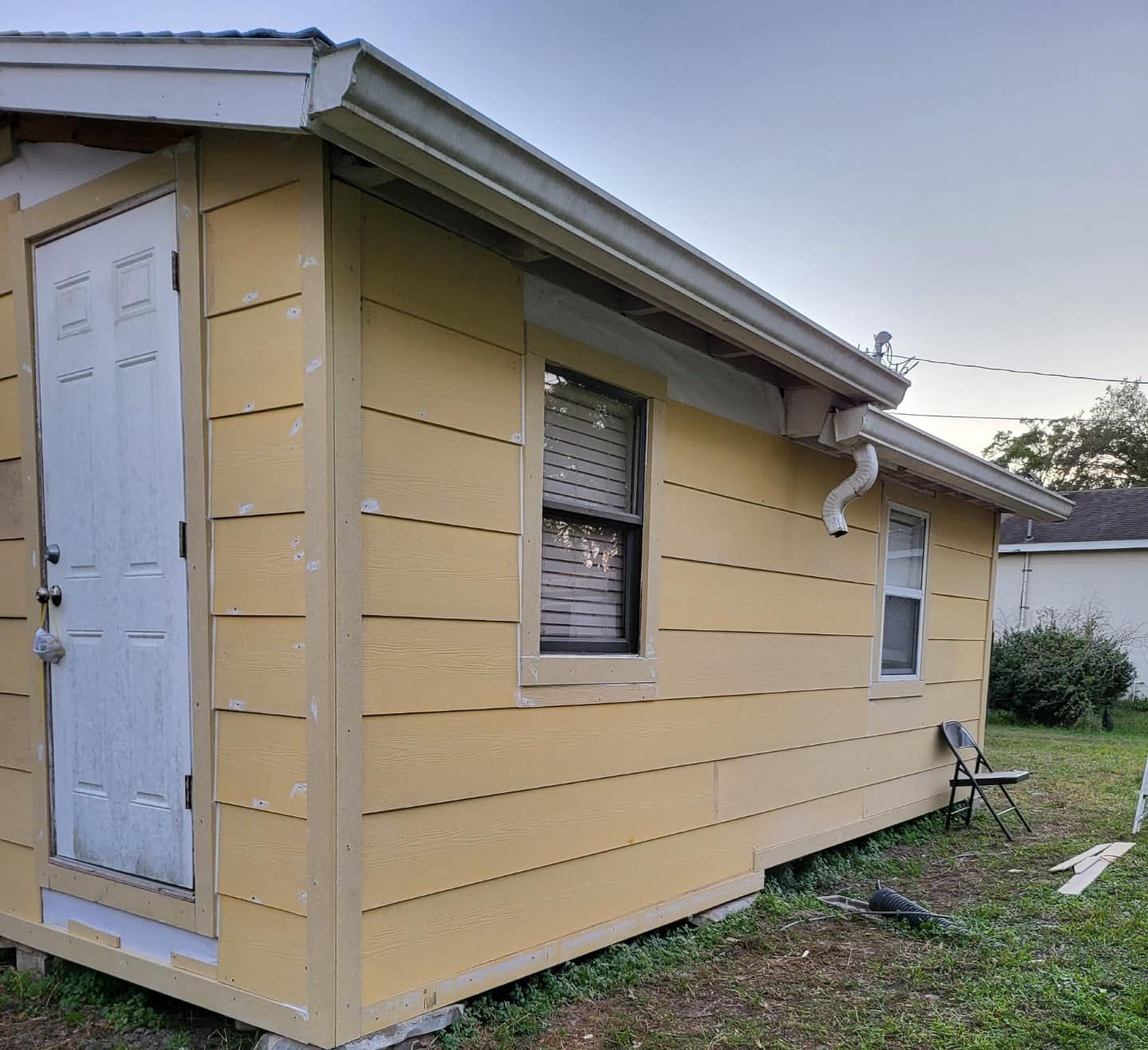 Yellow-sided small building with white door, windows, and trim, brown roof, and gutters, set on green grass.